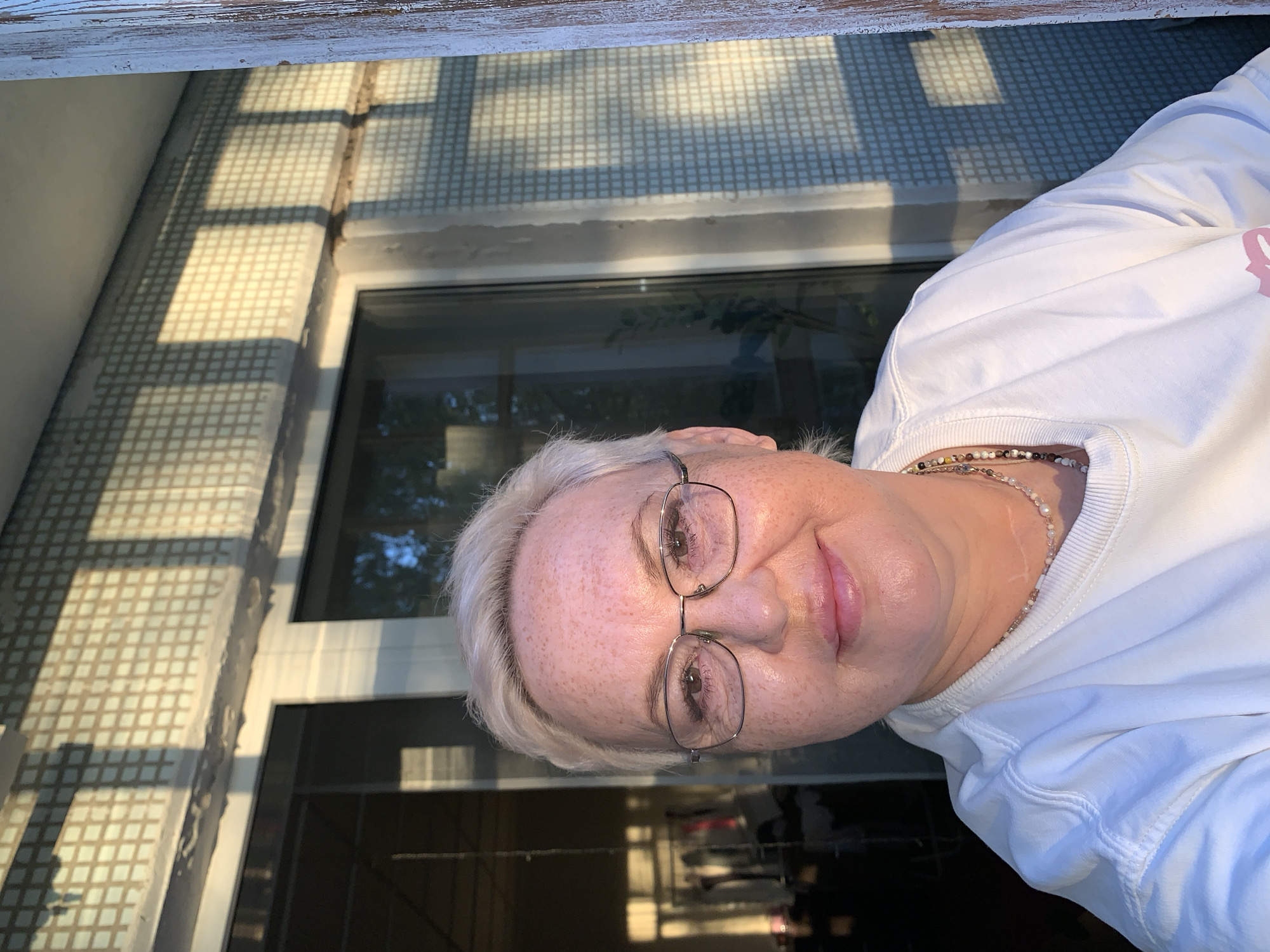 A middle-aged woman with short, light-colored hair and eyeglasses poses for a picture. She is wearing a white sweatshirt and a beaded necklace. She appears to be outdoors, adjacent to a building with a tiled exterior, possibly a windowsill. The building's architecture includes window features with reflections of trees. The lighting suggests it is daytime, with strong sunlight casting shadows on the tiled exterior. The specific location within the city of Chaikovsky, Russia, cannot be determined from the image.