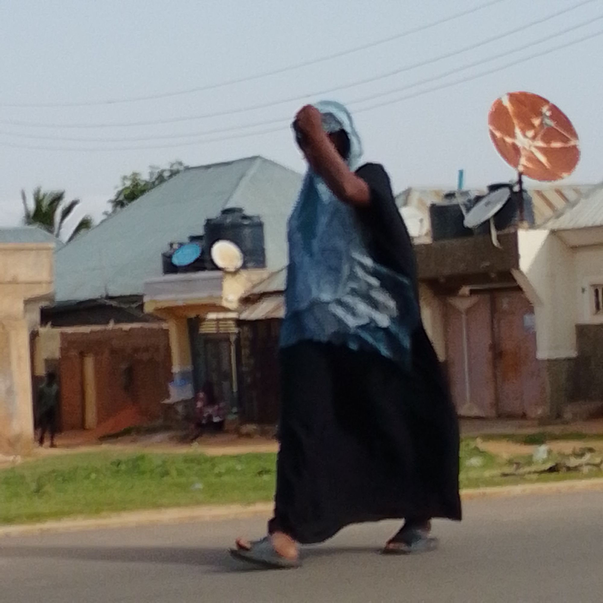 The image depicts a street scene in Gombe, Nigeria, likely during daytime with overcast skies. A person, appearing to be a woman due to the attire, is walking from left to right across the frame, partially obscuring a building in the background. She is wearing a dark, long skirt and a lighter blue garment draped over her shoulders and head, with her face covered by a light blue fabric. The building behind her has a light-colored, slanted roof, with a prominent orange satellite dish mounted on its side. Other satellite dishes, possibly smaller ones, are also visible on the roof. The foreground shows a paved road, with a strip of green grass bordering it to the left. Additional figures, less distinct, are present in the background, one standing near a building on the far left, and others near the building with the satellite dishes on the right. The environment appears to be a semi-urban or residential area, characterized by buildings and open ground.
