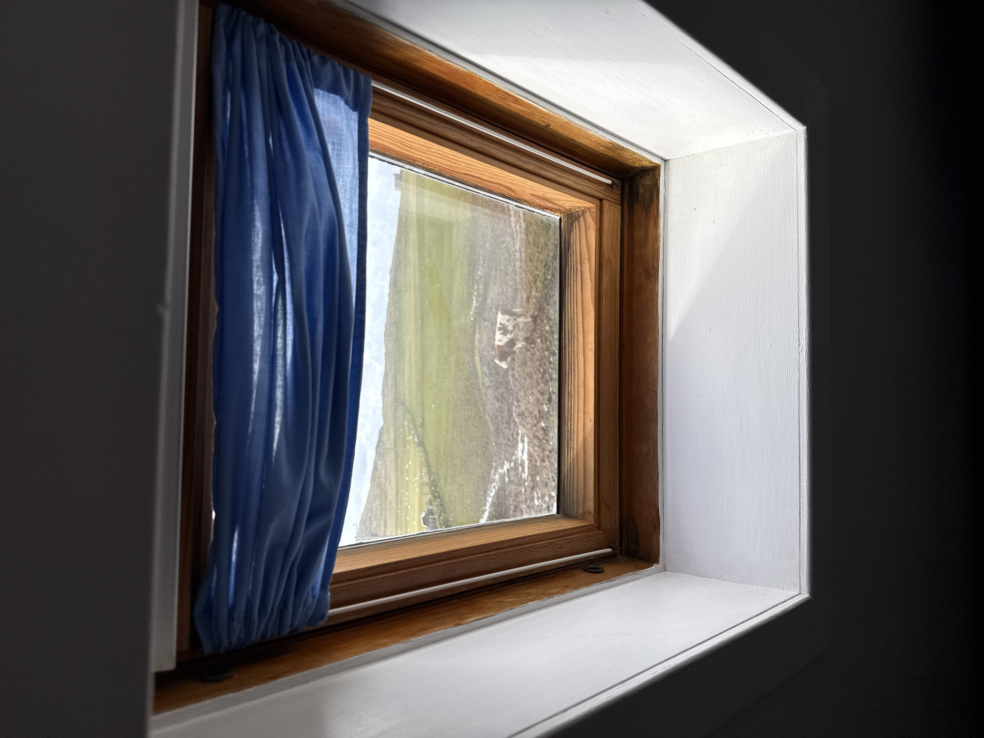 A wood-framed window, slightly open with a blue curtain partially covering the left side, is set into a white painted wall. The window offers a view of a green and brown landscape. The right side of the frame is heavily shadowed, suggesting a dim interior. The window appears to be situated within a room of Unknown, Unknown.