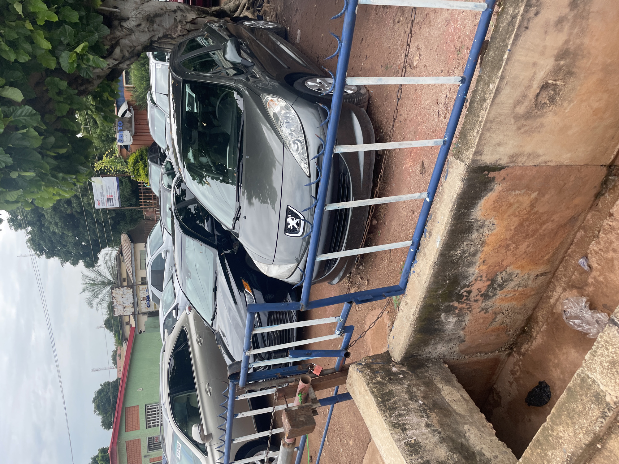 The image depicts an outdoor scene in Jos, Nigeria, featuring a row of parked cars behind a blue metal fence with white vertical bars. The cars visible include a dark-colored sedan in the foreground, a dark-colored hatchback to its left, and a silver SUV further back. A large tree with dense green foliage is on the right side of the frame, casting shadows. In the background, there are buildings, including one with a green facade and a red roof, and another with a lighter color. The ground is covered with reddish-brown earth and gravel. A concrete structure, possibly a drainage channel or a low wall, is in the foreground, with some debris visible inside it. The sky is overcast.