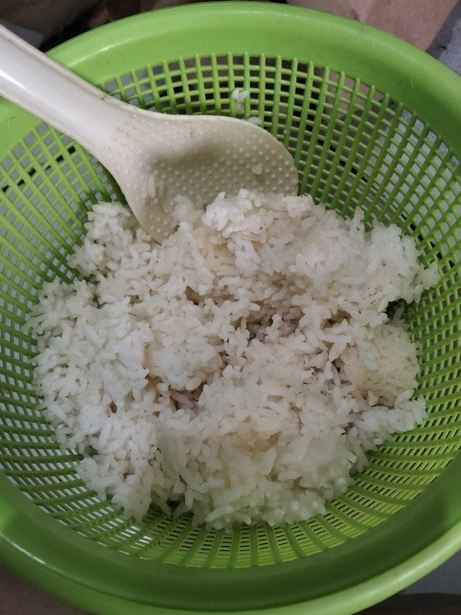 A top-down shot shows a green plastic colander filled with cooked white rice. A cream-colored rice paddle rests on top of the rice, its textured surface visible. The colander is placed on a brown surface. The scene is indoors, and the lighting is natural. The image does not provide any information about the location in Central Jakarta, Indonesia.