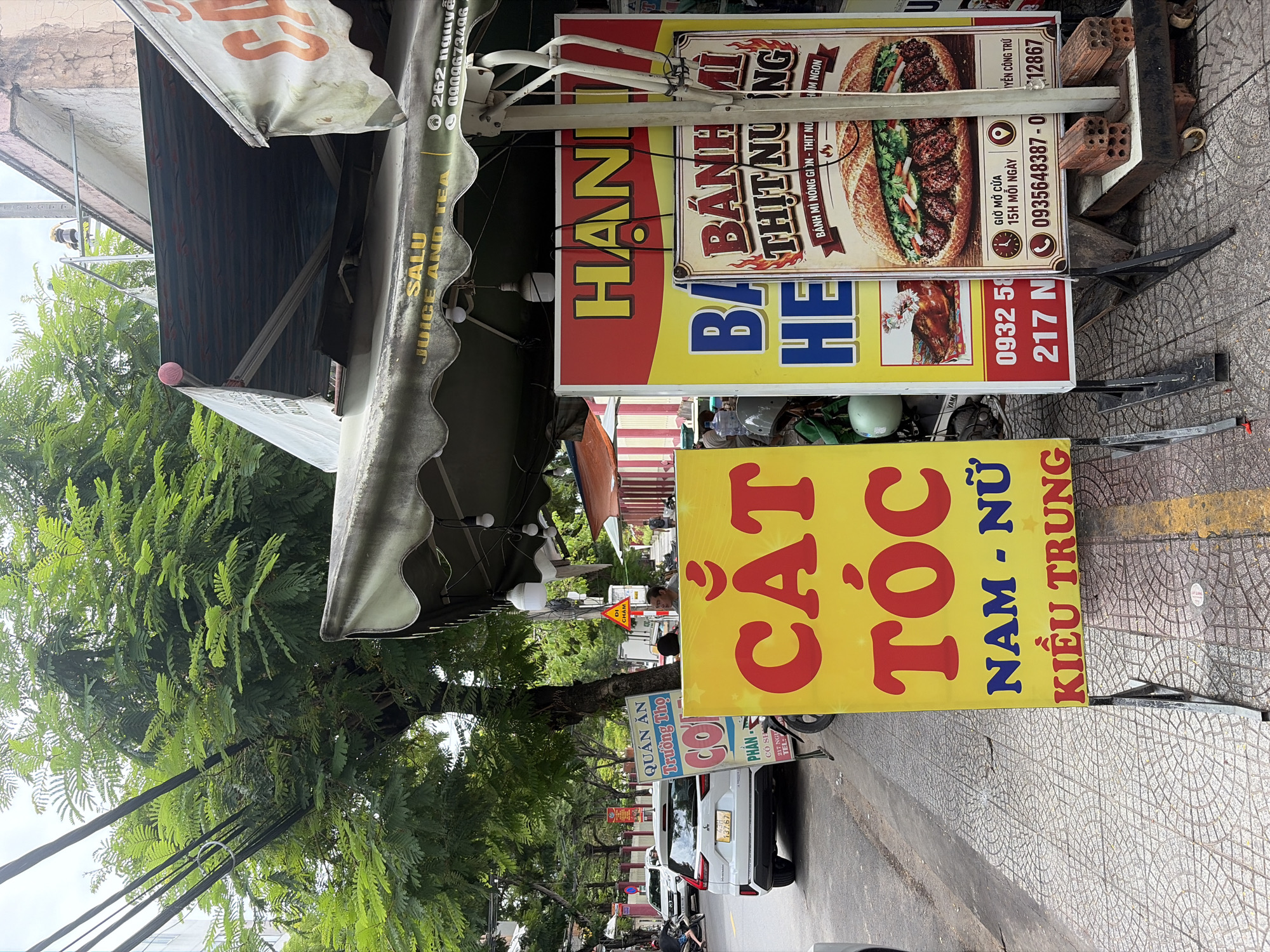 A street scene in Da Nang, Vietnam, features a sidewalk with various signs and businesses. In the foreground, a bright yellow sign with red text reads "CẮT TÓC NAM - NỮ KIỂU TRUNG," indicating a hair salon specializing in "Trung" (likely Chinese) hairstyles for men and women. To the right, a red and yellow sign advertises "BÁNH MÌ THỊT NƯỚNG" (grilled pork banh mi), with an image of the sandwich and details about opening hours and phone numbers. Another sign to the left of the banh mi advertisement, partially obscured, has blue text.
Adjacent to these signs, an awning extends over what appears to be a food stall or shop, with text indicating "SALU JUICE AND TEA." Further down the street, a white SUV with a Vietnamese license plate is parked, and people on motorcycles are visible in the background. Trees line the sidewalk, and buildings form the urban backdrop. The ground is paved with patterned tiles. The overall atmosphere suggests a bustling commercial area.