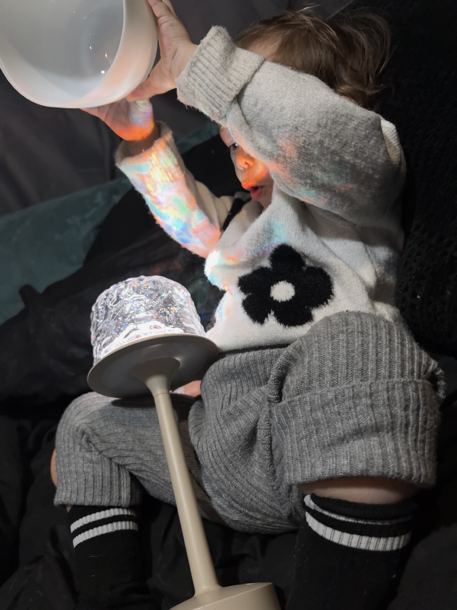 A young child sits on a dark surface, possibly a couch or bed, illuminated by a multi-colored light emanating from a nearby lamp. The child is wearing a white sweater with a black flower design, grey pants, and black socks with white stripes. The child is holding a white bowl above their head. The lamp has a clear, patterned shade and a beige base. The location is unknown, and there is no evidence to support that this image was taken in Noviy, Russia.