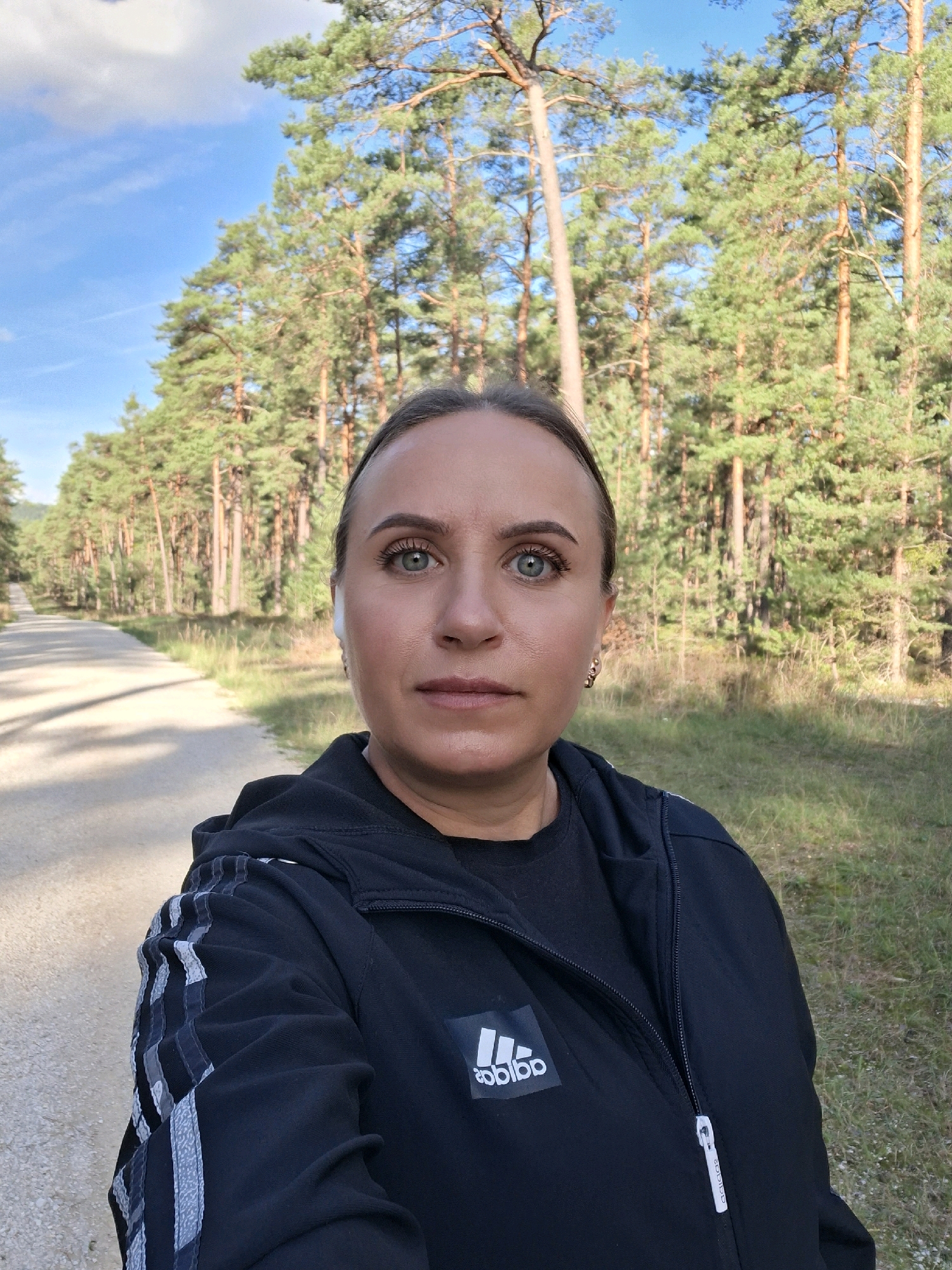 A woman with light skin and dark hair pulled back is facing the camera, wearing a black hooded jacket with grey and white stripes on the sleeves, and an Adidas logo on the left chest. She is wearing white wireless earbuds. The background shows a paved path, a grassy area, and a dense forest of tall pine trees under a blue sky with scattered clouds. The scene is outdoors, likely in a park or forest, given the path and trees. The lighting suggests it is daytime.