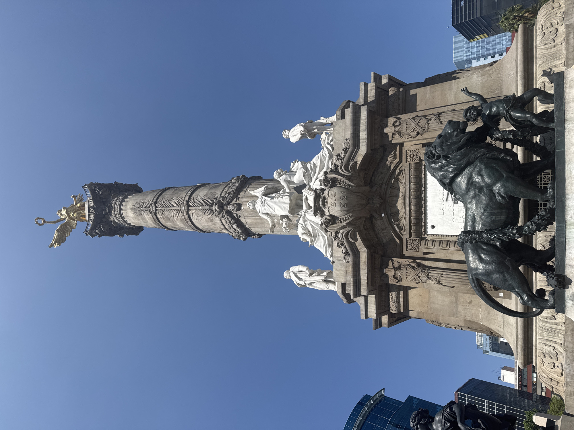 The image captures the Independence Monument in Mexico City, Mexico, viewed from a low angle. The monument consists of a tall column topped by a golden statue of the Winged Victory. The base of the column features several white marble statues. At the monument's base are bronze statues depicting a person struggling with a lion. The sky is clear and blue. Modern buildings can be seen in the background.