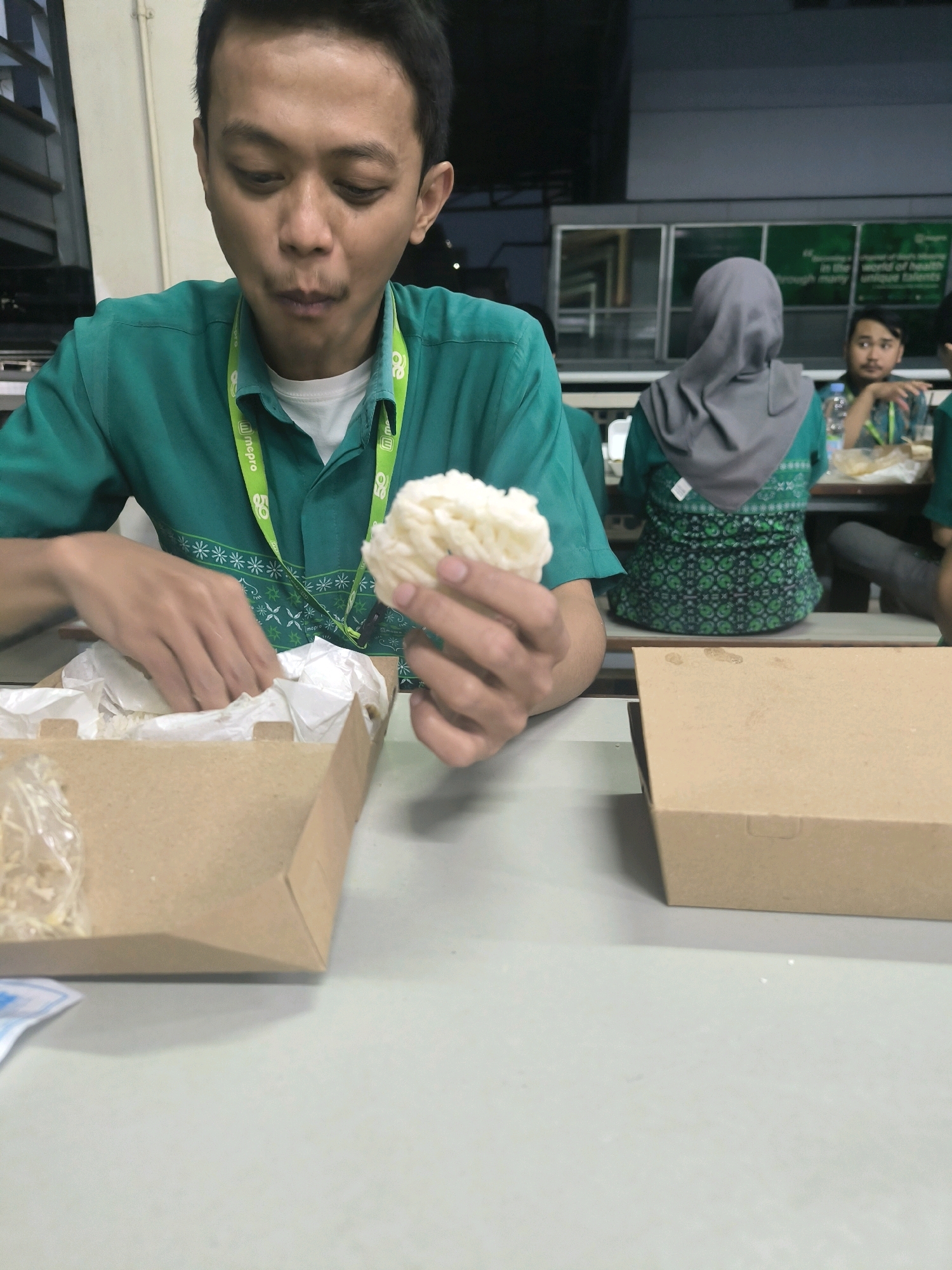 A medium shot shows a young man wearing a green shirt with a lanyard around his neck, holding a white puffed snack in his right hand and reaching into a brown paper food container with his left hand. The man is looking down at the food and appears to be eating. The food container, lined with white paper, is on a light gray table in front of him, next to a closed brown cardboard box. In the background, a woman wearing a grey hijab and a green patterned shirt is seated at a table. Further in the background, another person is partially visible. The environment suggests an indoor setting, possibly a cafeteria or dining area, with what looks like posters or signage on the wall in the far background. The overall scene depicts a casual mealtime.