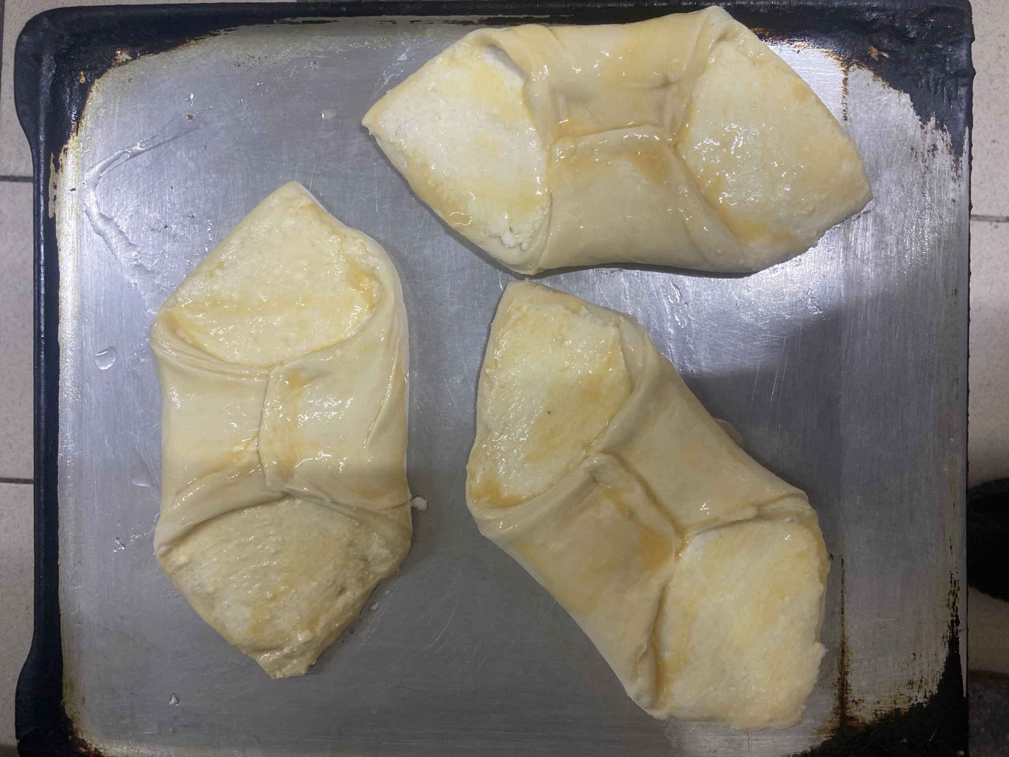 The image shows three raw pastries on a metal baking sheet. The pastries are light in color and appear to be brushed with a glaze, possibly egg wash. They are rectangular with pinched corners. The baking sheet is worn and has a dark, possibly burnt, residue along the edges. The pastries are arranged haphazardly on the sheet. The background appears to be a tiled surface.
The location of the image is not evident and cannot be determined from the image alone. The pastries appear to be homemade.