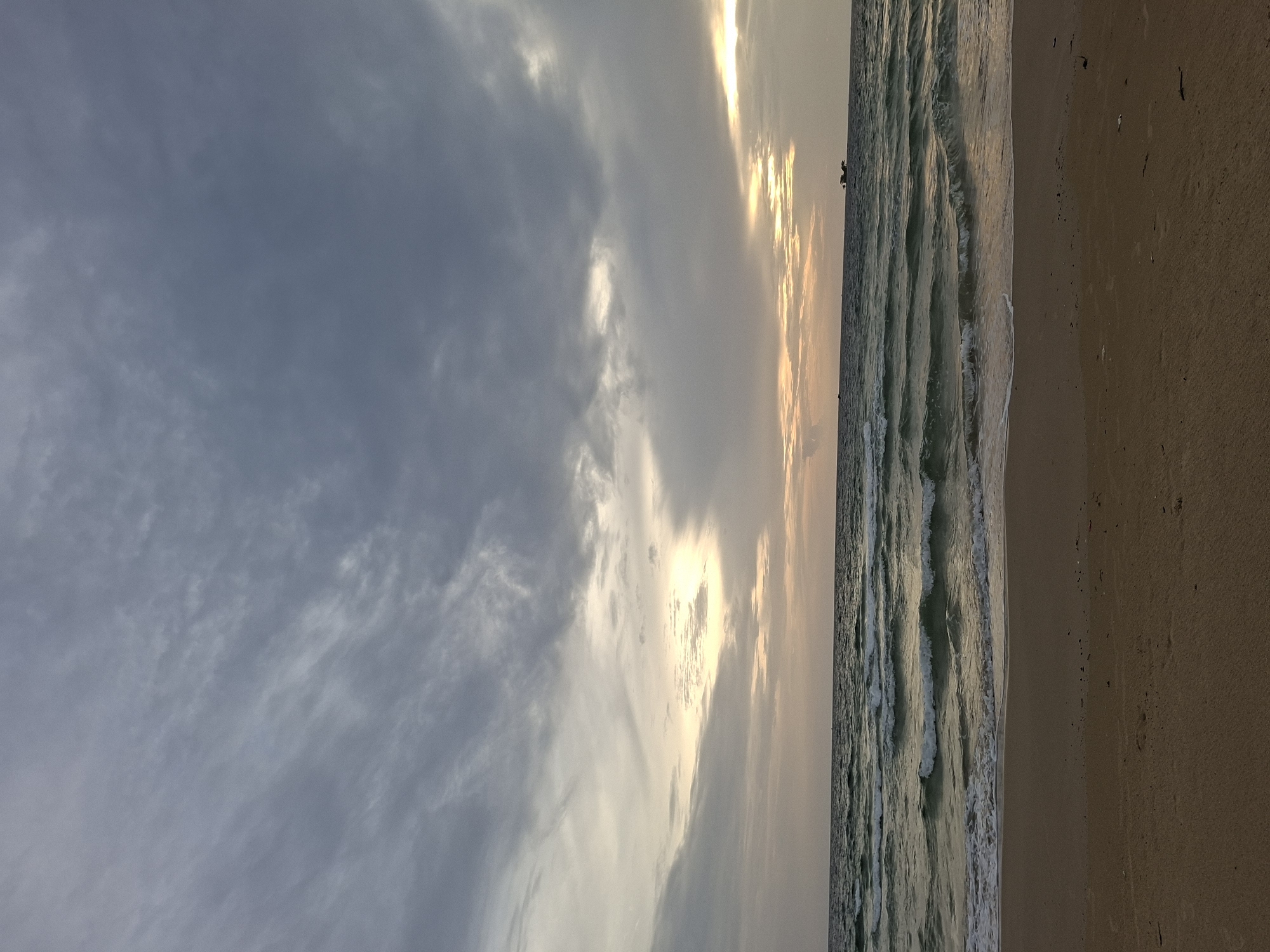 A naturalistic, eye-level, vertical shot captures a serene beach scene in Phú Quốc, Vietnam, under a dramatic sky. The horizon is divided into three distinct bands: the sandy shore in the foreground, the sea in the middle, and the sky above. The beach, composed of fine, light brown sand, is mostly empty, with a few faint footprints visible. The sea is a deep, tranquil blue-green, with small, white-capped waves gently lapping at the shore, creating a foamy white line where they meet the sand. A few distant objects are visible on the water, potentially small boats or buoys. The sky is a complex tapestry of clouds. In the upper half, dark grey clouds dominate, streaked with lighter grey and white, suggesting an approaching storm or the aftermath of one. However, a band of lighter, warmer-toned clouds near the horizon hints at a sunset or sunrise breaking through. This creates a striking contrast between the moody upper sky and the softer, more luminous lower sky. The overall atmosphere is one of peaceful solitude and the raw beauty of nature.