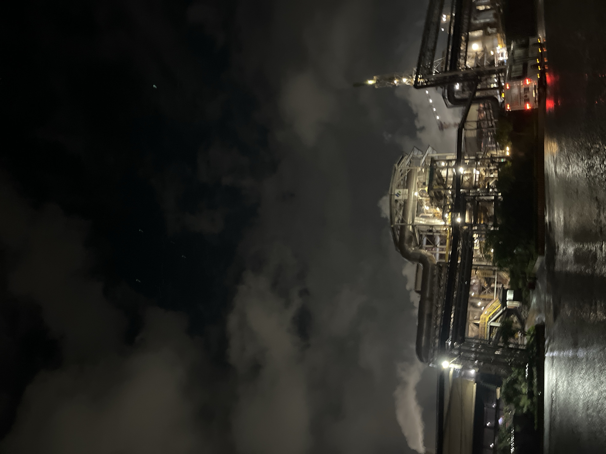 The image shows an industrial plant at night in Unknown, Indonesia. The facility is brightly illuminated with multiple structures connected by pipes and walkways. The ground appears wet, possibly from rain. The sky is dark and cloudy. There is a fire truck parked at the edge of the plant.