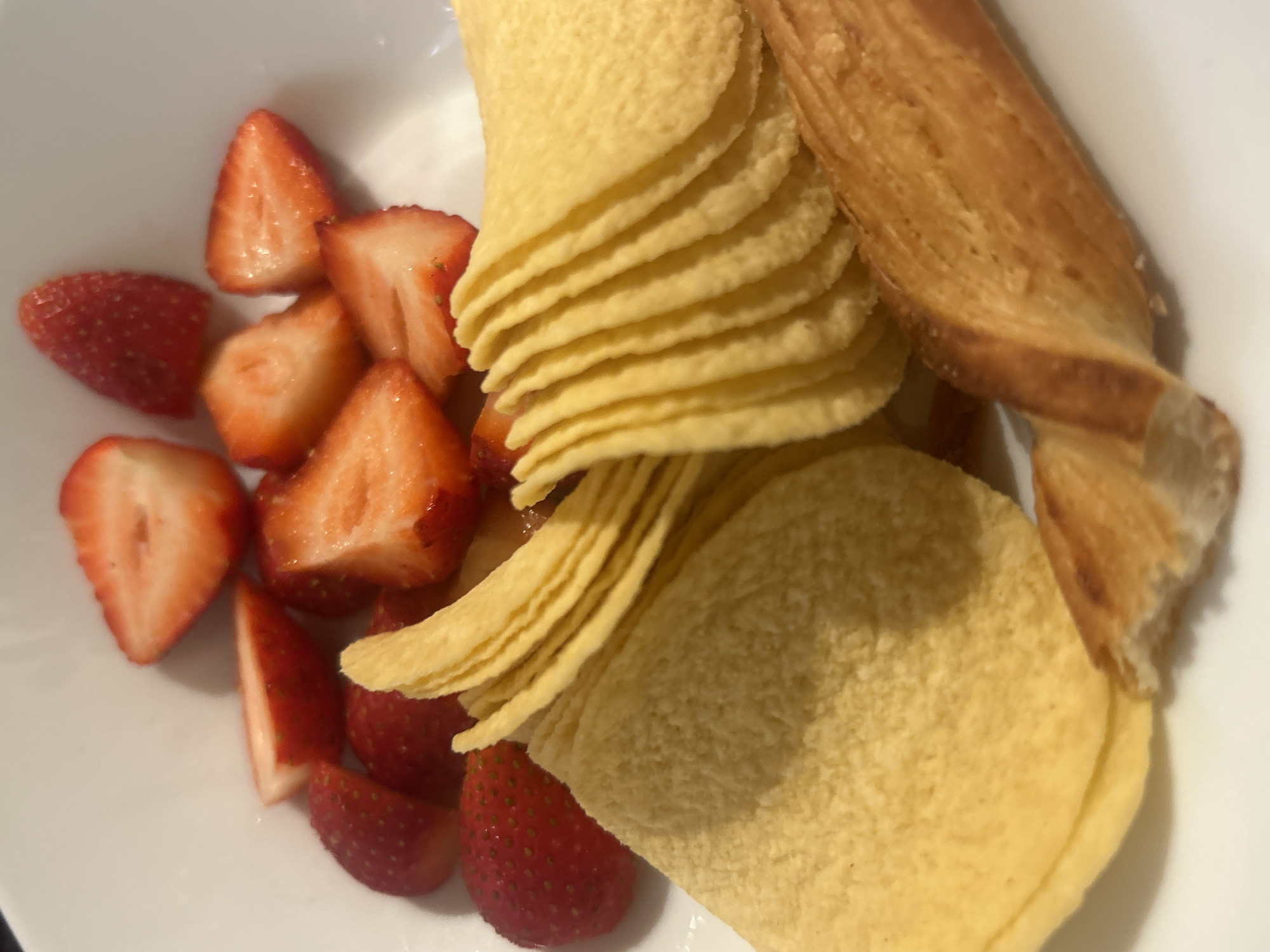 The image shows a white bowl containing a portion of sliced strawberries, a stack of ruffled potato chips, and a twisted breadstick. The strawberries are red and appear to be fresh. The potato chips are light yellow in color. The twisted breadstick has a golden-brown crust. The location is unspecified.