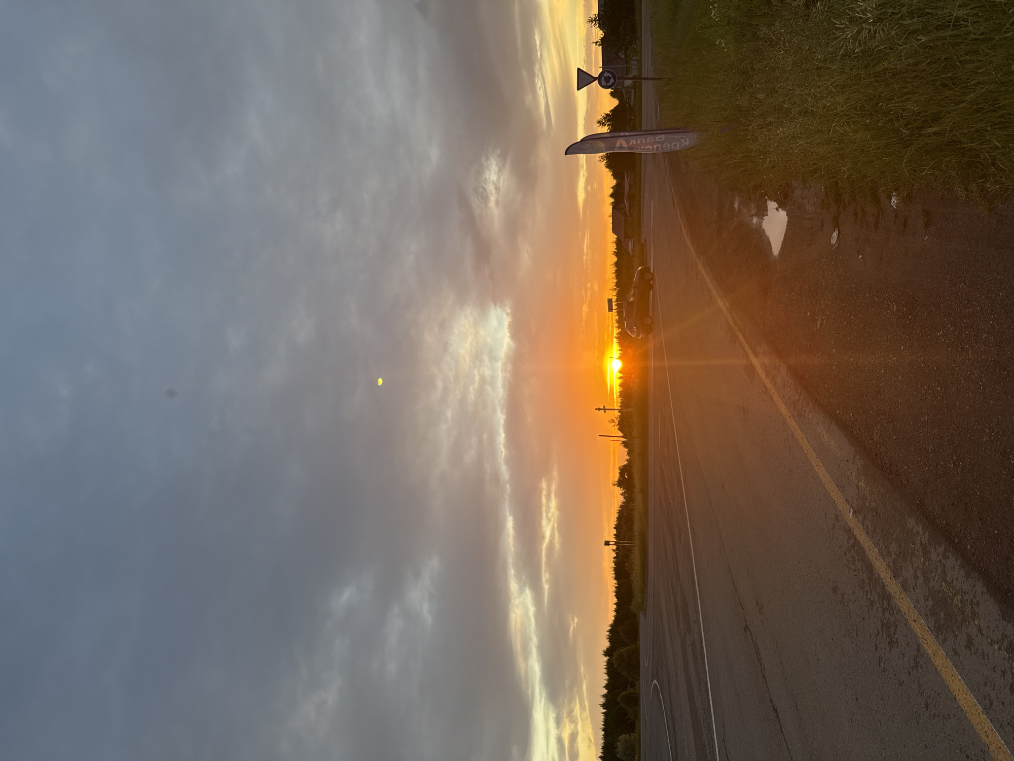 The image depicts a sunset over a paved road in Gol'yany, Russia. The sky is a dramatic blend of grey clouds on the left and a vibrant orange and yellow sunset on the right, with streaks of sunlight breaking through. The sun is low on the horizon, casting a warm glow.
On the right side of the road, there is lush green grass and a puddle of water reflecting the sky. Further down the road, a dark-colored car is parked. A sign with Russian text, "АВТОМОБИЛИ ЗАПЧАСТИ" (AUTOMOBILE SPARE PARTS), is visible on the side of the road. Two traffic signs are also present: a triangular sign with a red border and a circular sign indicating a roundabout.
The overall mood is serene and tranquil, characteristic of a sunset. The weather appears to be partly cloudy. The presence of the road and traffic signs suggests a semi-rural or suburban setting. The time of day is clearly sunset due to the prominent sun and its warm, low-angled light.