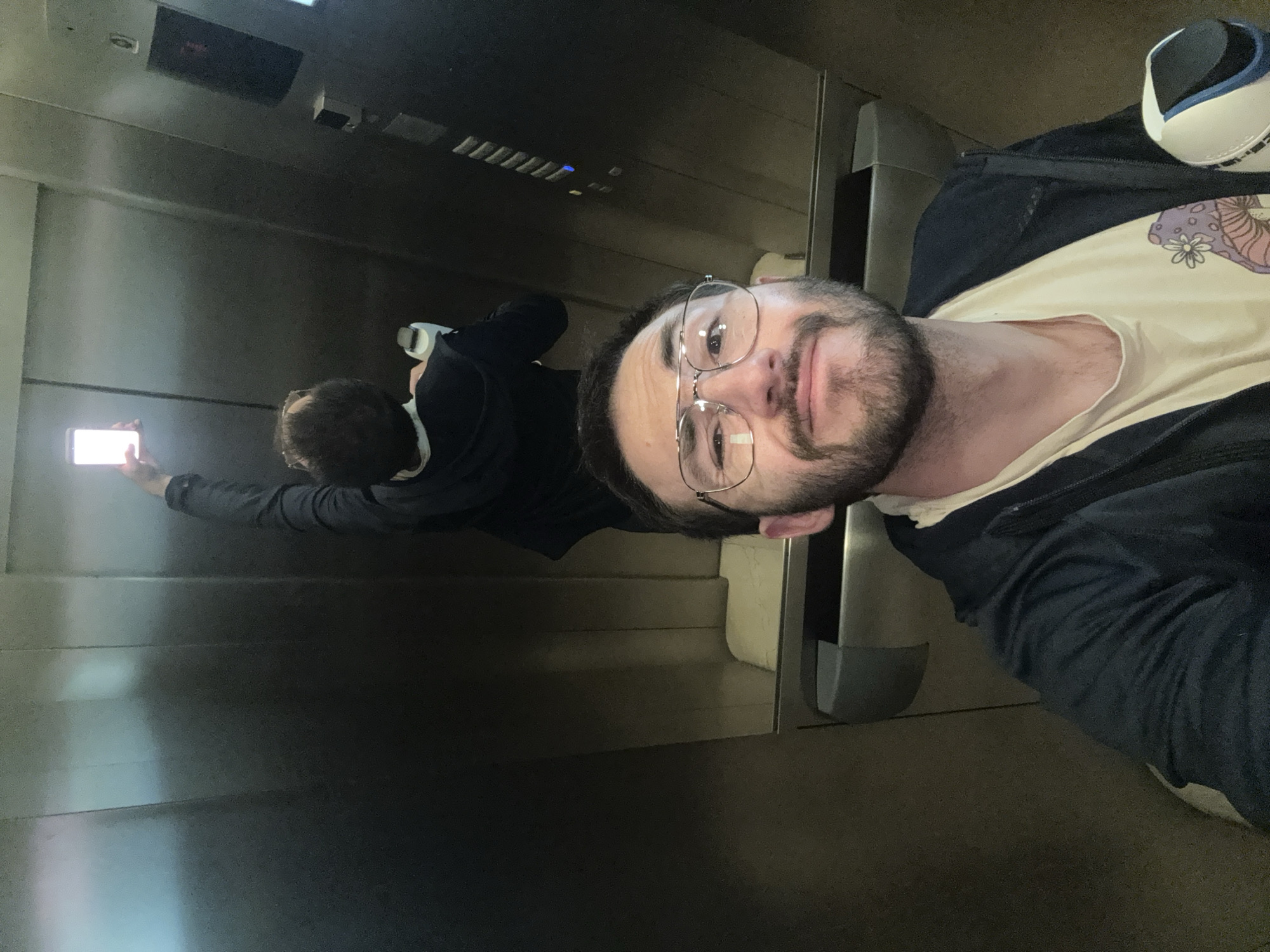 An indoor selfie photograph, rotated 90 degrees clockwise, captures a man inside an elevator in Huixquilucan de Degollado, Mexico. The man, positioned in the foreground, features a beard, mustache, and glasses, with short, dark hair. He is dressed in a light-colored t-shirt, possibly cream or light yellow, adorned with a purple and white floral or mushroom graphic on the left chest area, and a dark (likely black or navy blue) zip-up jacket over it. He looks directly at the camera with a subtle, calm expression.
Further back in the elevator, a reflection of a person, or possibly the same man, is visible on the polished metallic wall. This reflected figure has dark hair and wears a dark long-sleeved top, holding a smartphone with its screen brightly illuminated and pointing it upwards. The glowing phone screen acts as a strong light source in the background.
The elevator's interior is characterized by reflective metallic walls. A portion of a white and blue athletic shoe can be seen near the foreground man's shoulder. On the far left of the image, small red lights, likely from an elevator control panel or floor indicator, are faintly visible in the reflection. The lighting within the elevator is moderate, with the phone's light being the most intense. The image documents a casual moment, likely a selfie being taken. No discernible text is visible other than the small indicator lights.