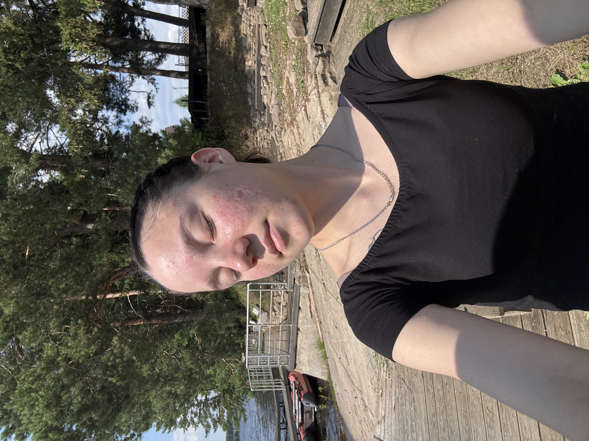 A young woman with her eyes closed is taking a selfie outdoors, likely in Pozdyshki, Russia, given the presence of a lake, pine trees, and wooden boardwalks. She has dark hair pulled back, and her face shows signs of acne. She is wearing a black t-shirt and a silver chain necklace. The background features a body of water with a small red boat docked, a wooden pier with railings, and a dense forest of pine trees under a blue sky with scattered clouds. The ground is a mix of wooden planks and rocky terrain with sparse green vegetation. The lighting suggests it is a sunny day.