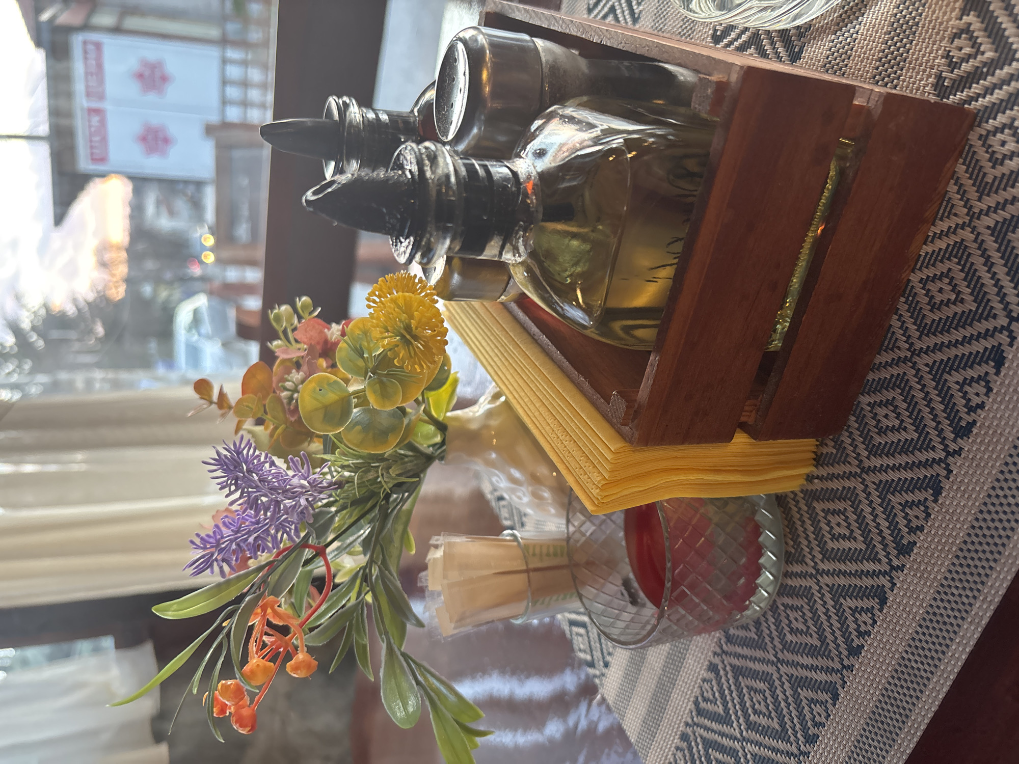 A close-up shot shows a table setting in what appears to be a restaurant, with a focus on a wooden container holding oil and vinegar bottles, yellow napkins, and a small vase of artificial flowers. The setting is on a table with a patterned tablecloth in shades of blue and white. In the background, visible through a window, there is an out-of-focus view of a street scene with signs in what appears to be Cyrillic script, suggesting an Eastern European location. Based on the location information given, this scene is in Sapareva Banya, Bulgaria. No people are visible in the shot.