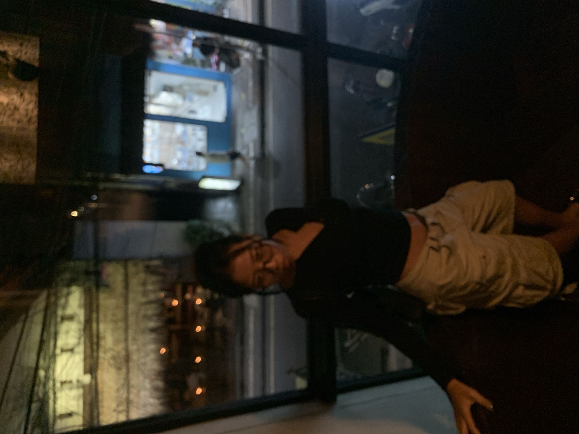 A medium, indoor shot, taken inside a building, with a person leaning forward and looking out of a window. The window offers a view of what appears to be the street lights of Ubud District, Indonesia. The person is wearing glasses, a black shirt with long sleeves, and beige shorts. They are leaning on a dark surface, presumably the window sill, and their hand is visible near the bottom right corner of the image. A building is visible in the distance through the window. The overall lighting is dim, with the brightest areas being the lights outside the window.