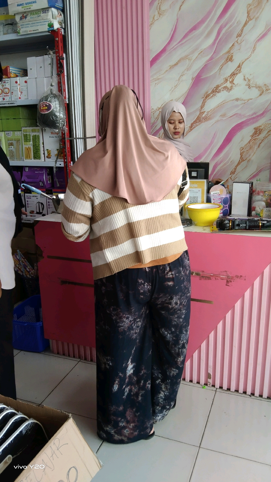 The image depicts an indoor scene, possibly a store, with two individuals visible. The primary subject is a person wearing a beige hijab and a striped sweater over dark, patterned pants. They are facing away from the camera, looking towards a counter. Behind them, another person, also wearing a hijab, is partially visible, looking down at something on the counter.
The environment features a pink counter with various items displayed on it, including a yellow bowl, a laptop, and other miscellaneous objects. Behind the counter, a wall is decorated with a pink and white marble-like pattern. To the left of the main subject, shelves are stocked with products, including what appear to be food items in boxes. A blue basket is visible on the floor to the left.
The flooring is tiled with a light color. A portion of a cardboard box with writing is visible in the lower left corner, with the text "vivo Y20" and what appears to be handwritten Indonesian text. The scene suggests a retail environment in a commercial area, potentially in Karawang, Indonesia, given the context of the request.