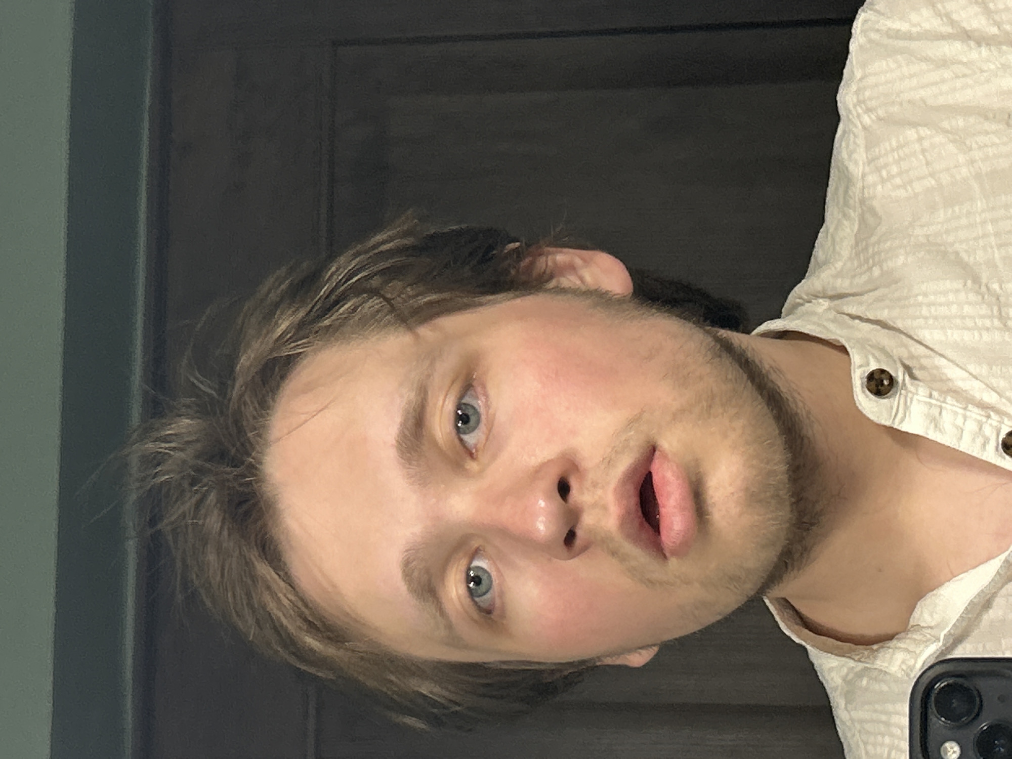 A close-up, off-kilter portrait shows a Caucasian male with light brown hair and blue eyes. He has stubble on his face and is wearing a light-colored shirt with brown buttons. Part of a black smartphone is visible in the bottom right corner. The background consists of what appears to be a dark door and a gray-green wall. The location is Unknown, Unknown.