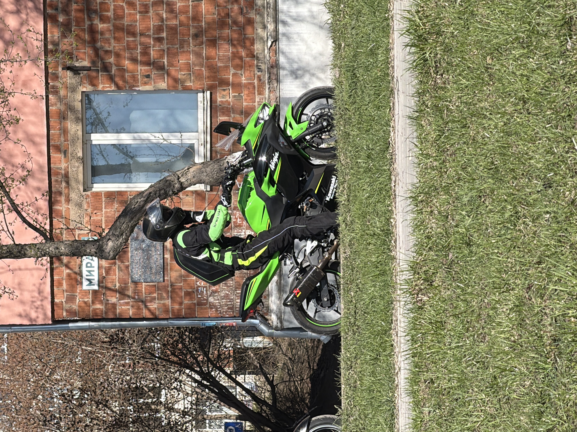 A green and black Kawasaki Ninja motorcycle is parked on a city sidewalk in Votkinsk, Russia, next to a grassy area. The rider, dressed in black and green motorcycle gear with a helmet, is on the bike. A brick building with a window and a sign reading "МИРА" in Cyrillic is in the background. A tree branch extends across the window and the motorcycle. The scene is viewed from a high vantage point.