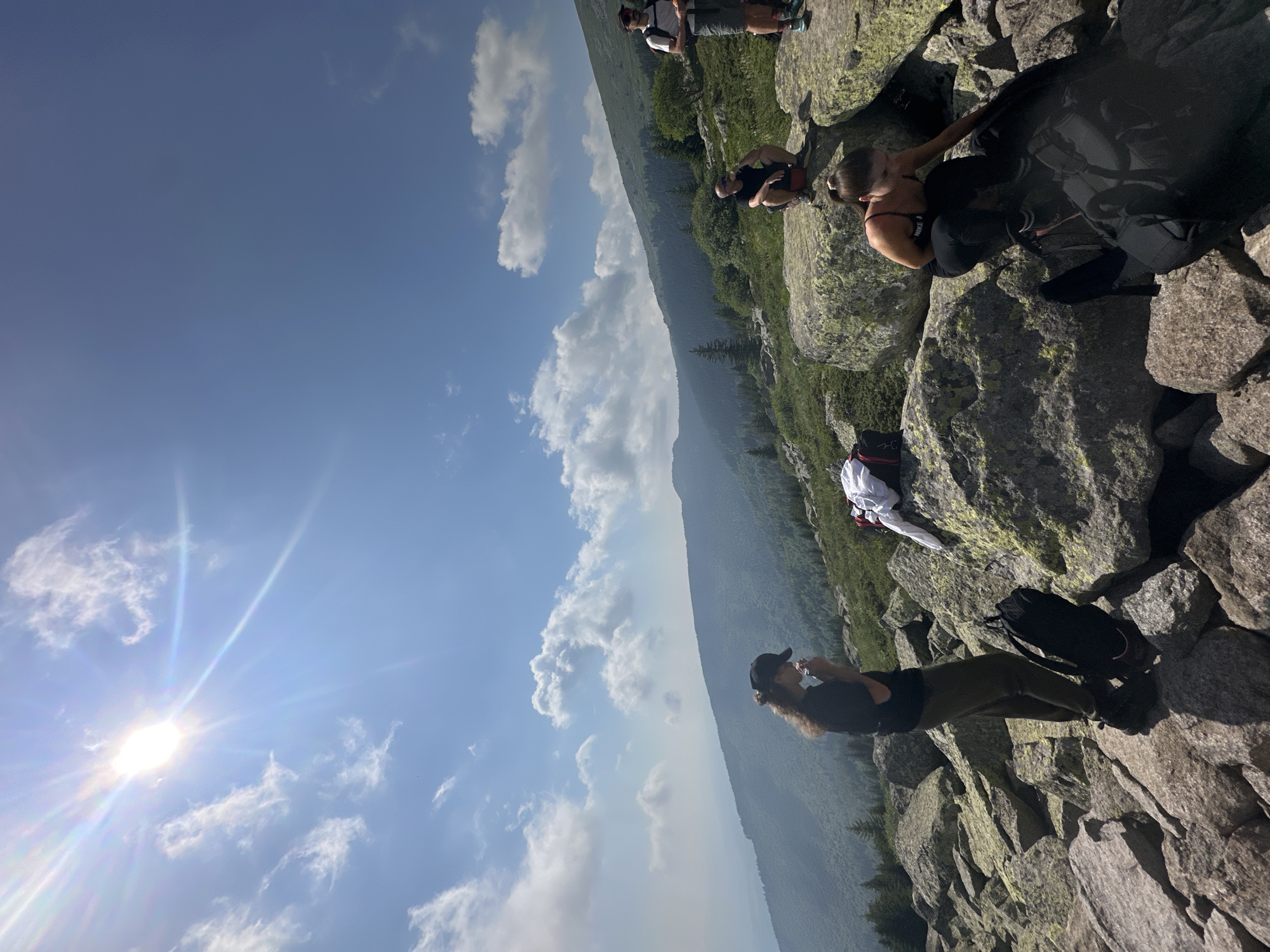 The image depicts a group of people on a rocky, mountainous terrain, likely on Vitosha Mountain, near Bistritsa, Bulgaria. The sun is bright, with visible lens flare, and there are scattered clouds in the bright blue sky.
In the foreground, a woman with long, curly hair and a black baseball cap is standing on large rocks, holding something to her mouth. She is wearing a black t-shirt and olive-green pants. A black backpack is positioned next to her.
To the right of the standing woman, a woman with her hair in a ponytail is crouching on a large rock, reaching towards something out of frame. She is wearing a black tank top with "NIKE" visible and black leggings. Another black backpack is in front of her.
Further to the right and slightly behind, a man is crouched on a rock, holding a bottle to his lips, wearing a black t-shirt and shorts. Another man is standing to the far right, wearing a backpack and shorts, with another person partially visible behind him.
The environment is characterized by rugged, grey and green-tinged rocks, with sparse green vegetation and trees growing on the slopes in the background. The landscape stretches out into a hazy, mountainous vista with forested valleys. Several backpacks and articles of clothing are scattered amongst the rocks, suggesting a rest stop during a hike.