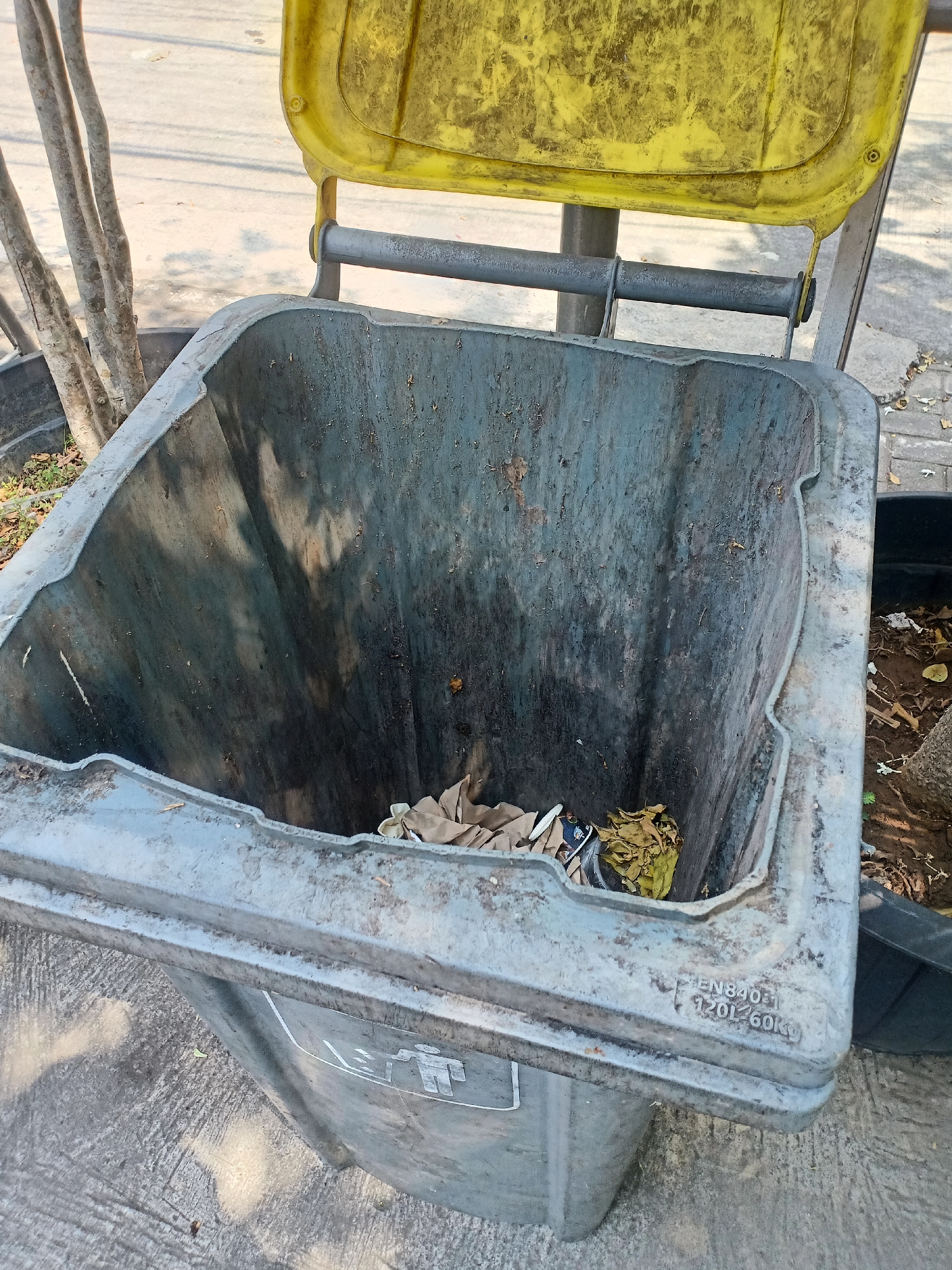 An open, weathered, gray plastic trash bin is shown outdoors, with its yellow lid partially open, revealing the interior of the bin. The bin is empty except for a small amount of dry leaves and some scattered paper. The bin has a rough texture and appears to be made of durable plastic. The interior walls are stained and marked from previous use. The bin is situated on a paved surface, likely a sidewalk or pathway, with trees and other vegetation visible in the background. A portion of a black plastic container is also visible to the right of the trash bin. The scene is lit by natural sunlight, casting shadows on the ground and the bin. The inscription "EN840-1 120L/60KG" is visible on the exterior of the bin, indicating its capacity and standard compliance. The context suggests this is a public waste receptacle in an urban setting, possibly in Semarang, Indonesia, given the presence of tropical vegetation and outdoor paving.