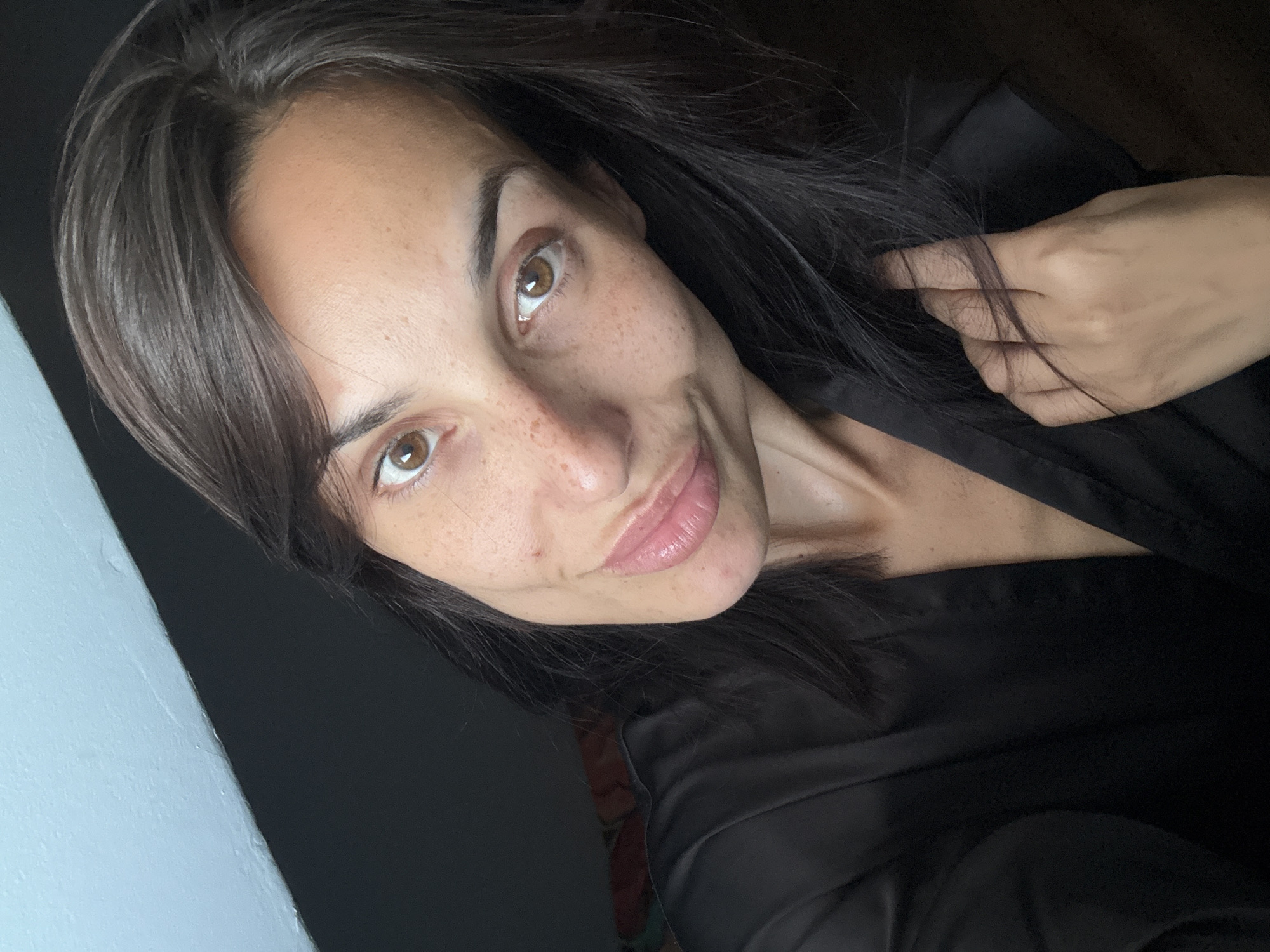 The image is a close-up, selfie-style photograph of a woman with dark hair and brown eyes. She is wearing a black satin robe. The background is dark, with a visible sliver of a light blue surface at the top left, suggesting an indoor setting, possibly a bedroom, with a sloped ceiling or wall. The woman has freckles across her nose and cheeks, and her lips are slightly parted. Her right hand is raised and resting near her hair. There is no indication of the city of Pleven, Bulgaria, in the image.