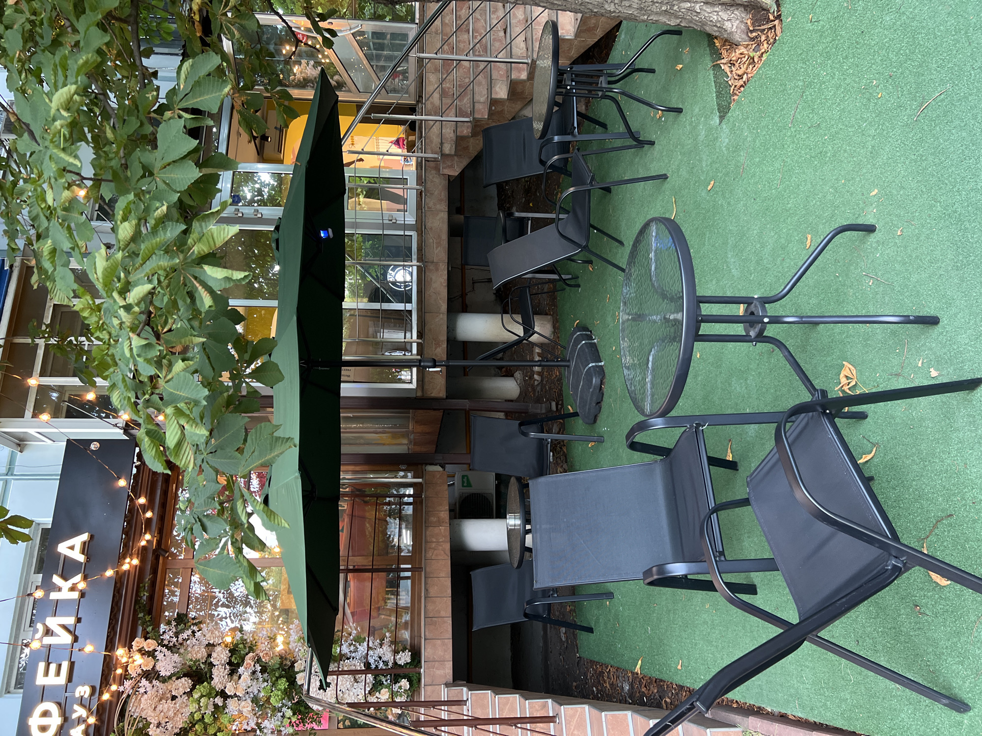 An outdoor seating area of a cafe is depicted in Krasnodar, Russia. The area is covered with green artificial turf and furnished with black metal chairs and glass-topped tables. A large dark green umbrella is set up, providing shade. In the background, the cafe's facade is visible, featuring a sign with white letters and decorative string lights. A staircase leads up to the cafe entrance. Lush green foliage from a tree overhangs the seating area. The overall atmosphere is calm and inviting, suggesting a relaxed urban setting.