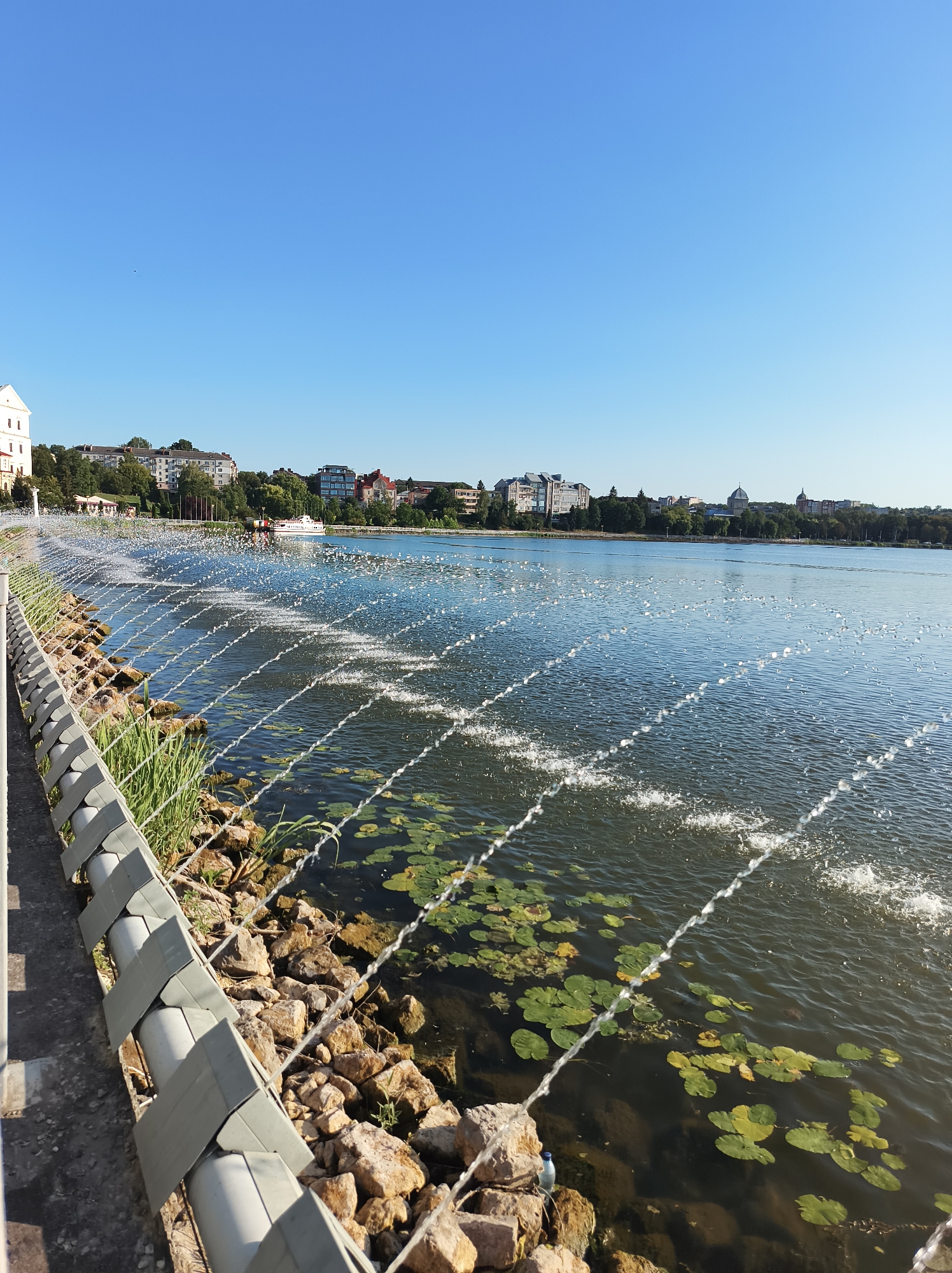 The image depicts the Ternopil pond in Ternopil, Ukraine, on a clear, sunny day. The foreground features a promenade with a decorative metal railing along the edge of the pond. Adjacent to the promenade, the pond's edge is reinforced with large rocks and greenery. Numerous water jets create a mesmerizing pattern of spraying water across the surface of the pond, forming a distinct line of fountains. Lily pads dot the water's surface, indicating a natural ecosystem within the urban setting. In the background, the city skyline rises, characterized by a mix of residential buildings, some with visible windows and balconies, and trees. A white boat with passengers is visible on the water, adding a sense of activity. The sky is a clear, vibrant blue, characteristic of a pleasant summer day.
