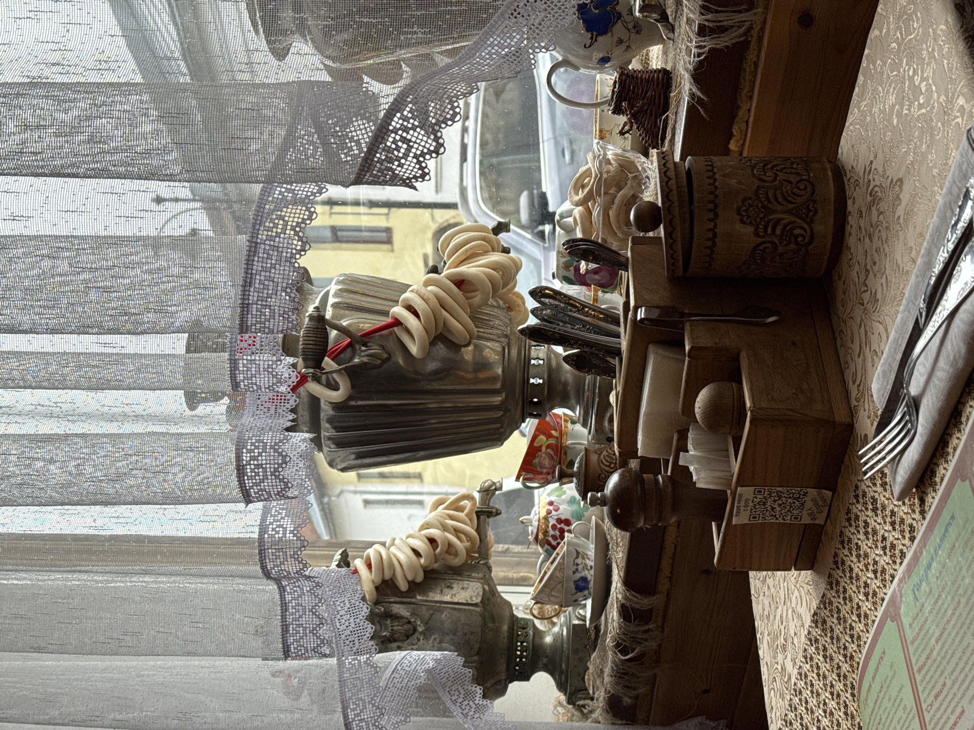 The image captures a cozy interior scene, likely a restaurant or cafe in Ново-Никольское, Russia, viewed through a window. The main subjects are two traditional samovars, intricately decorated and featuring strings of sushki (ring-shaped bread) wrapped around them. One samovar is larger and in the foreground, while the other is partially visible behind it.
The setting appears to be a dining area with a wooden table on the right, covered by a patterned, light-brown tablecloth. On the table, a silver fork rests on a folded grey napkin. Adjacent is an open menu or booklet with green and brown sections. A wooden condiment holder, featuring a small QR code with text "Наше меню с фото" (Our menu with photos), "English", "Español", and "中国人", is also on the table. Other visible text on the menu includes "Медовухо гос", "Медовуха Монастырском" (Monastery Mead), and "Сэт Медов" (Mead Set). Above the table, a carved wooden container is visible.
The window, which makes up the left side of the image, is covered by translucent white curtains with delicate lace edges, diffusing the daylight. Beyond the window, a blurry street scene with parts of a vehicle and buildings can be discerned, indicating it is daytime. Small, decorative teacups and pottery pieces are interspersed among the samovars and on shelves, contributing to a traditional and rustic atmosphere. No specific activity or event is actively taking place, but the setup suggests a dining experience.