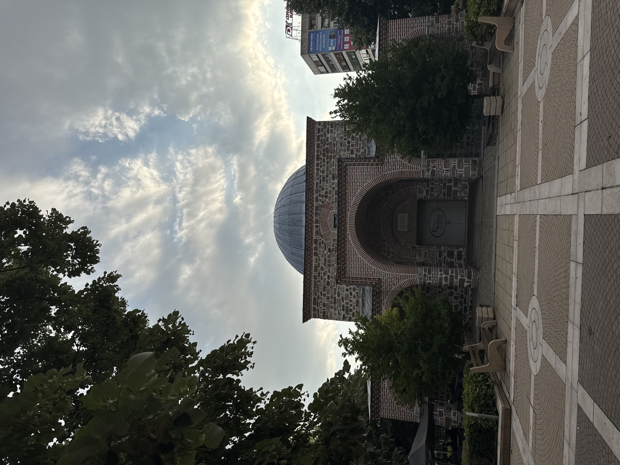 This is a photograph of the Bezisten (covered market) in Yambol, Bulgaria. The building features a large dome and is constructed with stone and brick. In front of the building, there is a paved courtyard with benches. Trees are visible on either side of the building and in the foreground. In the background, a modern apartment building with advertisements can be seen. The sky is cloudy.