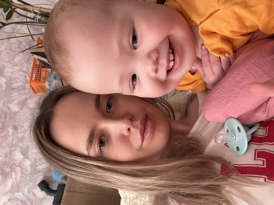 A woman and baby pose together in a selfie. The woman, with light brown hair, lies on a floral-patterned surface. A smiling baby, wearing an orange top, nestles close to her, its face the most prominent part of the image. The baby's smile reveals several teeth. A pink fabric and a light blue pacifier are nearby. A glimpse of a plant in a basket, along with some objects, is visible in the background.