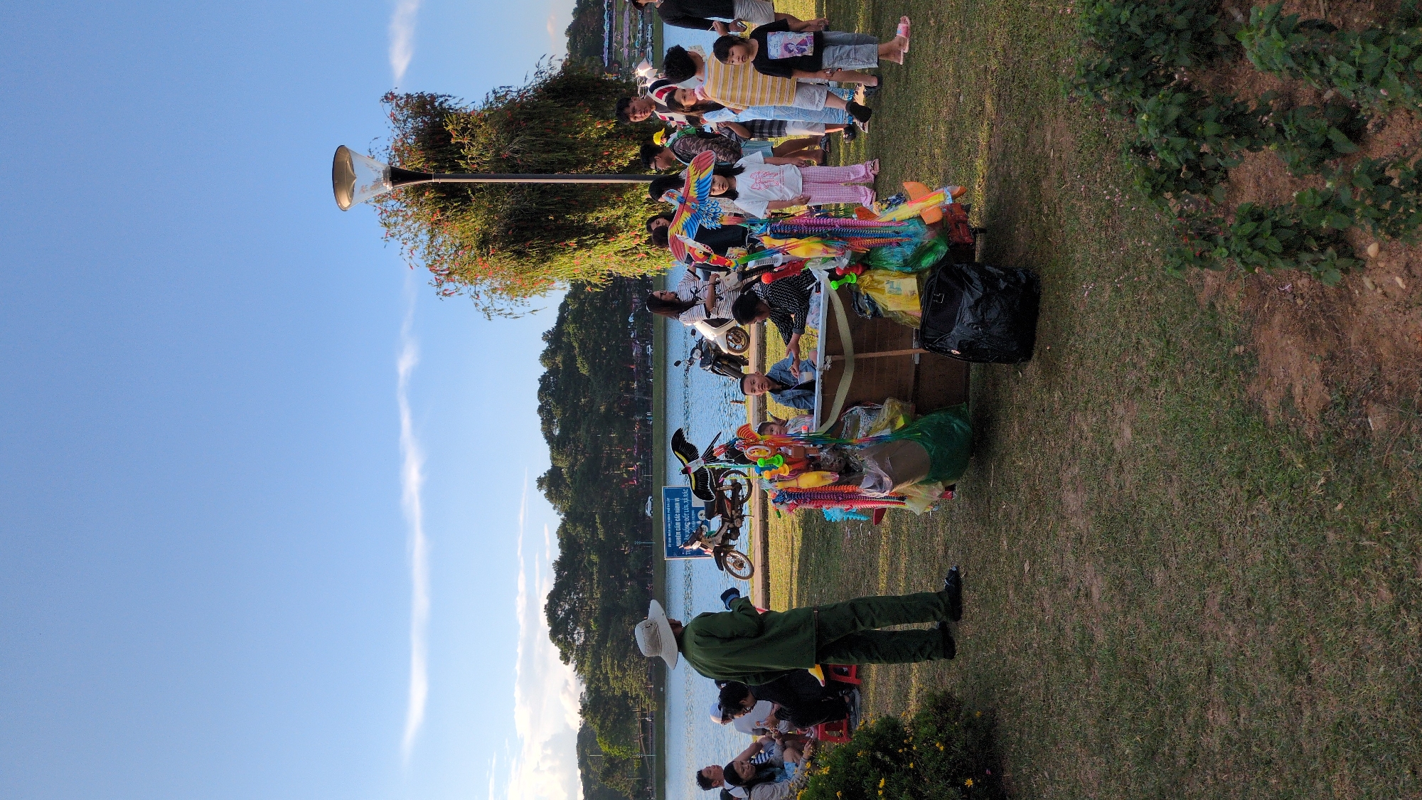 The image depicts a bustling scene at Xuan Huong Lake in Da Lat, Vietnam, during the late afternoon. The foreground features a colorful stall selling various toys and trinkets, including kites, plastic swords, and stuffed animals, attracting a group of children and adults. A man in a green uniform and hat, possibly a park ranger or vendor, stands near the stall, observing the surroundings. In the background, the serene expanse of Xuan Huong Lake is visible, with lush green trees lining its shores and mountains in the distance. Several motorbikes are parked near the lake, indicating a popular recreational spot. The sky is a clear blue, with a few wispy clouds, suggesting pleasant weather. The overall atmosphere is lively and familial, with people enjoying the outdoor environment and the array of goods on offer.