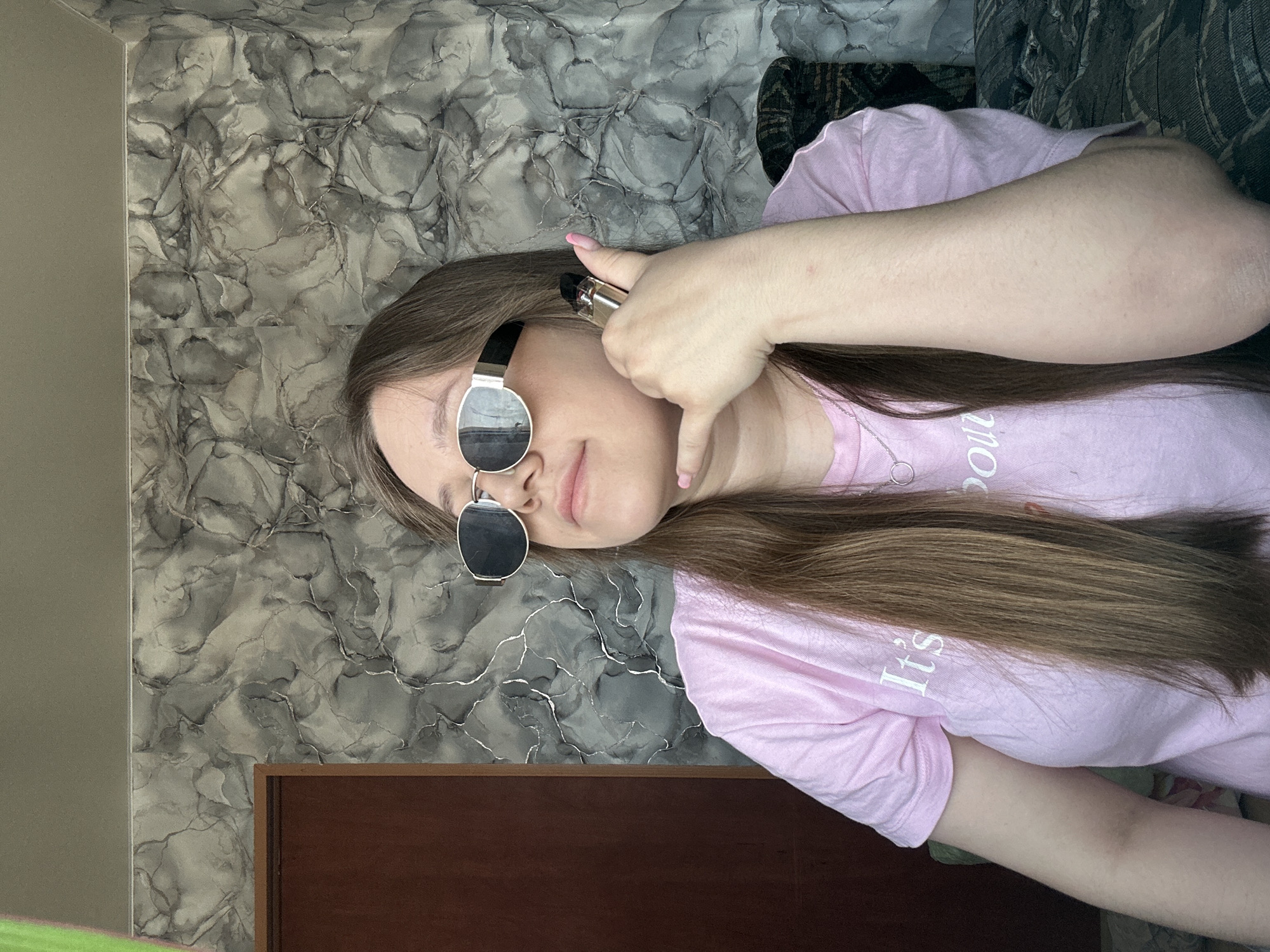 A woman lies sideways on an upholstered surface in what appears to be an indoor environment. She wears sunglasses with reflective lenses, a light pink T-shirt, and a silver necklace. Her left hand, with light pink nail polish, holds an object near her head, gesturing as if she is making a phone call. The background includes a wall with a marble-like pattern and a wooden piece. There is no evidence to confirm that the picture was taken in the city of Ольховка, Russia.