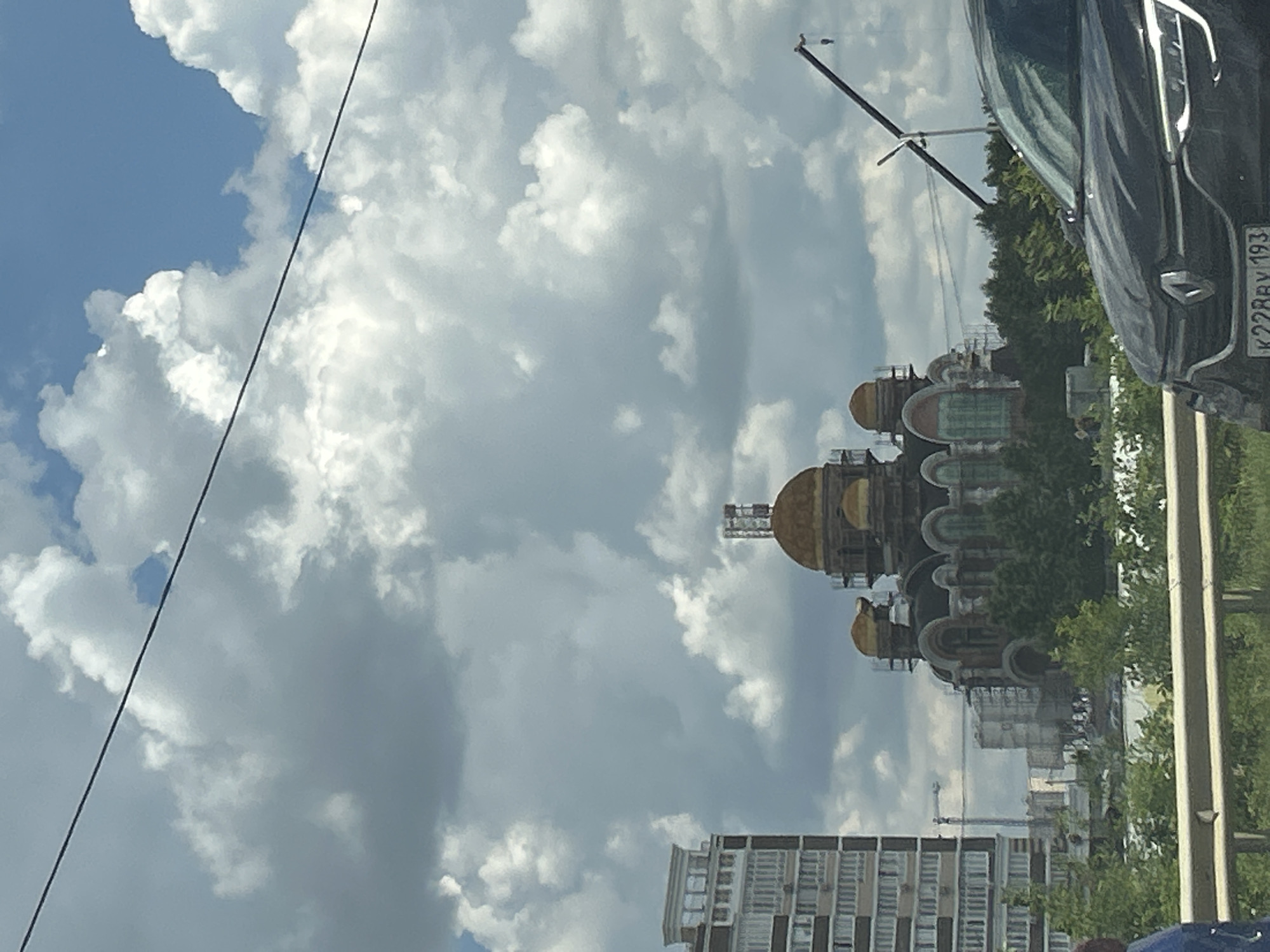 The image depicts an outdoor scene under a cloudy sky, with a strong sense of perspective due to the rotation of the image. The primary focus is on a distinctive building with multiple golden domes, suggesting a religious or historical architectural style, possibly under construction or renovation, as scaffolding is visible. Several other buildings, including a modern, multi-story residential block, are in the background. A portion of a dark-colored vehicle, possibly a car, is visible in the foreground on the right side, with a license plate displaying the text "K228BY 193". The presence of trees and a utility pole with wires contributes to the urban or suburban setting. The lighting suggests it is daytime, with the clouds creating a diffused, overcast light. The overall impression is one of a developing or established urban landscape in Russia, with a blend of old and new architecture.