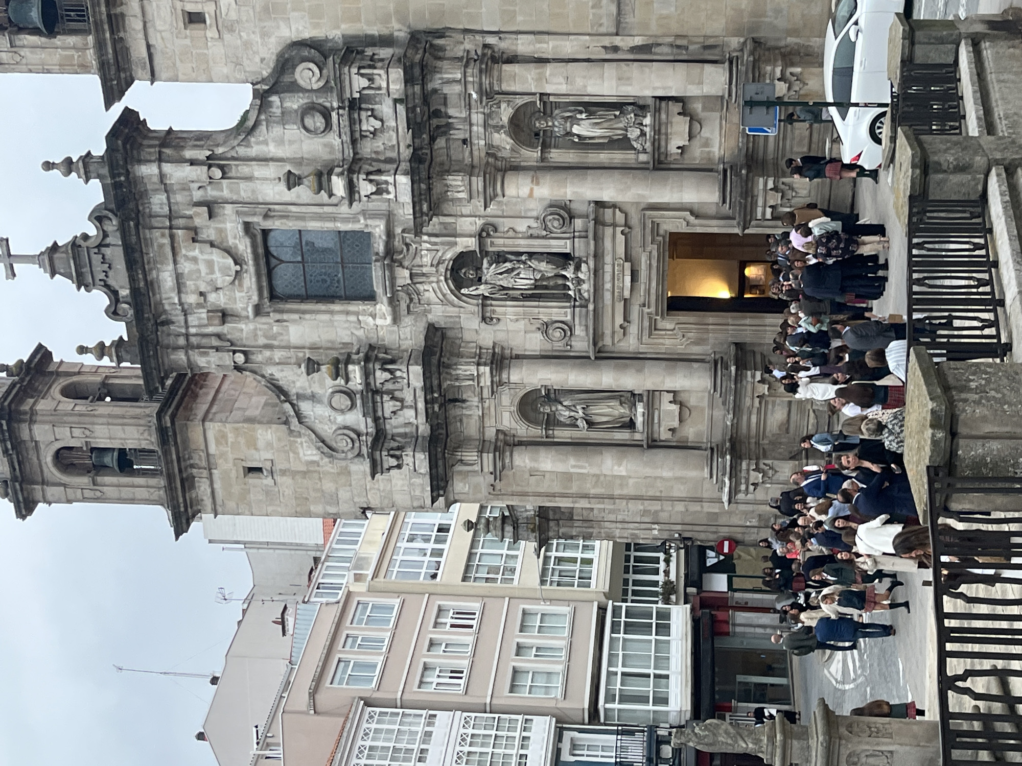 The image shows a stone building with ornate architectural details in A Coruña, Spain. The building appears to be a church, with statues in niches and a bell tower. A crowd of people is gathered in front of the building, many are in school uniforms and are on the left of the building. A white car is parked on the street nearby. Other buildings are visible in the background. The sky is overcast.
