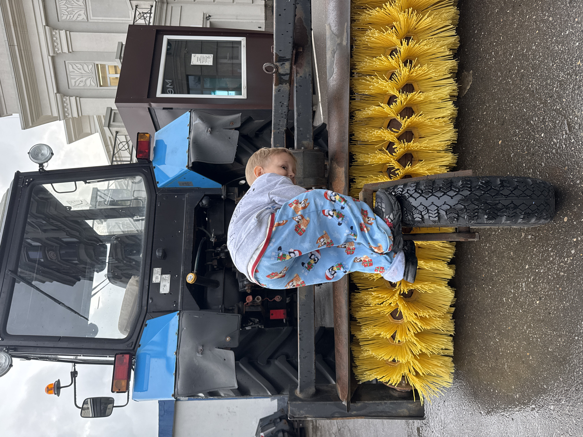The image shows a toddler in blue pajamas with a penguin print, sitting on a wheel attached to a large street sweeping machine. The toddler is leaning forward, looking down towards the yellow brushes of the machine. The street sweeper is blue and black, with a large tire and various attachments. In the background, a white building with classical architectural elements is visible, along with a dark brown kiosk with a glass window. The ground is wet, indicating it might be raining or has recently rained. There are no other people clearly visible in the image. The setting appears to be an outdoor urban environment, possibly a street or a maintenance yard in Moscow, Russia, given the building's architecture and the presence of the street sweeper.