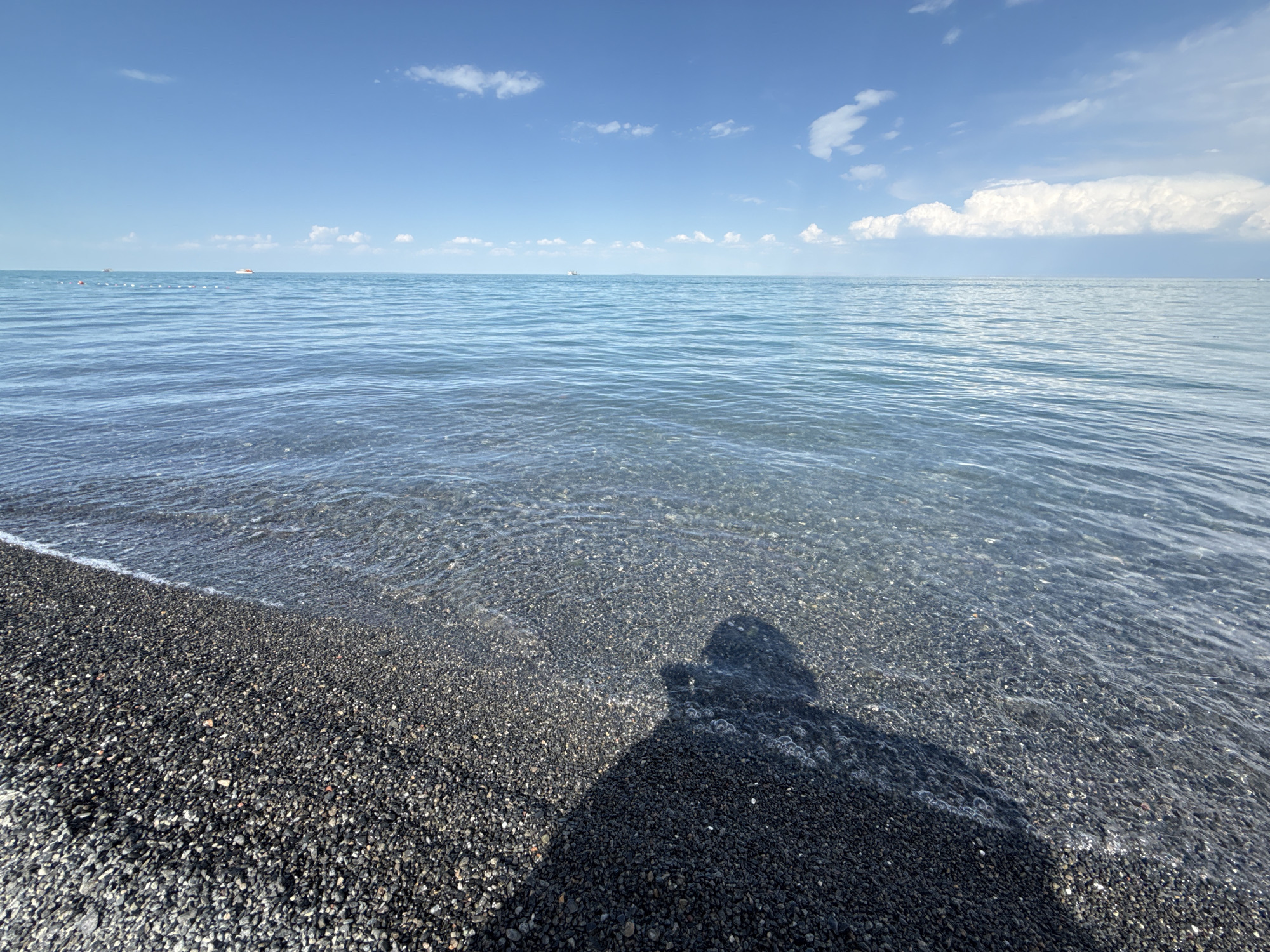 A wide shot captures a pebbled shoreline meeting the calm waters of a large lake under a clear blue sky dotted with white clouds. The water's surface reflects the sky and has a gentle ripple. In the distance, several small boats and buoys are visible on the horizon. A prominent shadow of a person holding a camera is cast across the pebbles and shallow water on the right side of the frame. The environment appears to be a tranquil beach scene in the city of Ақши, Kazakhstan.
