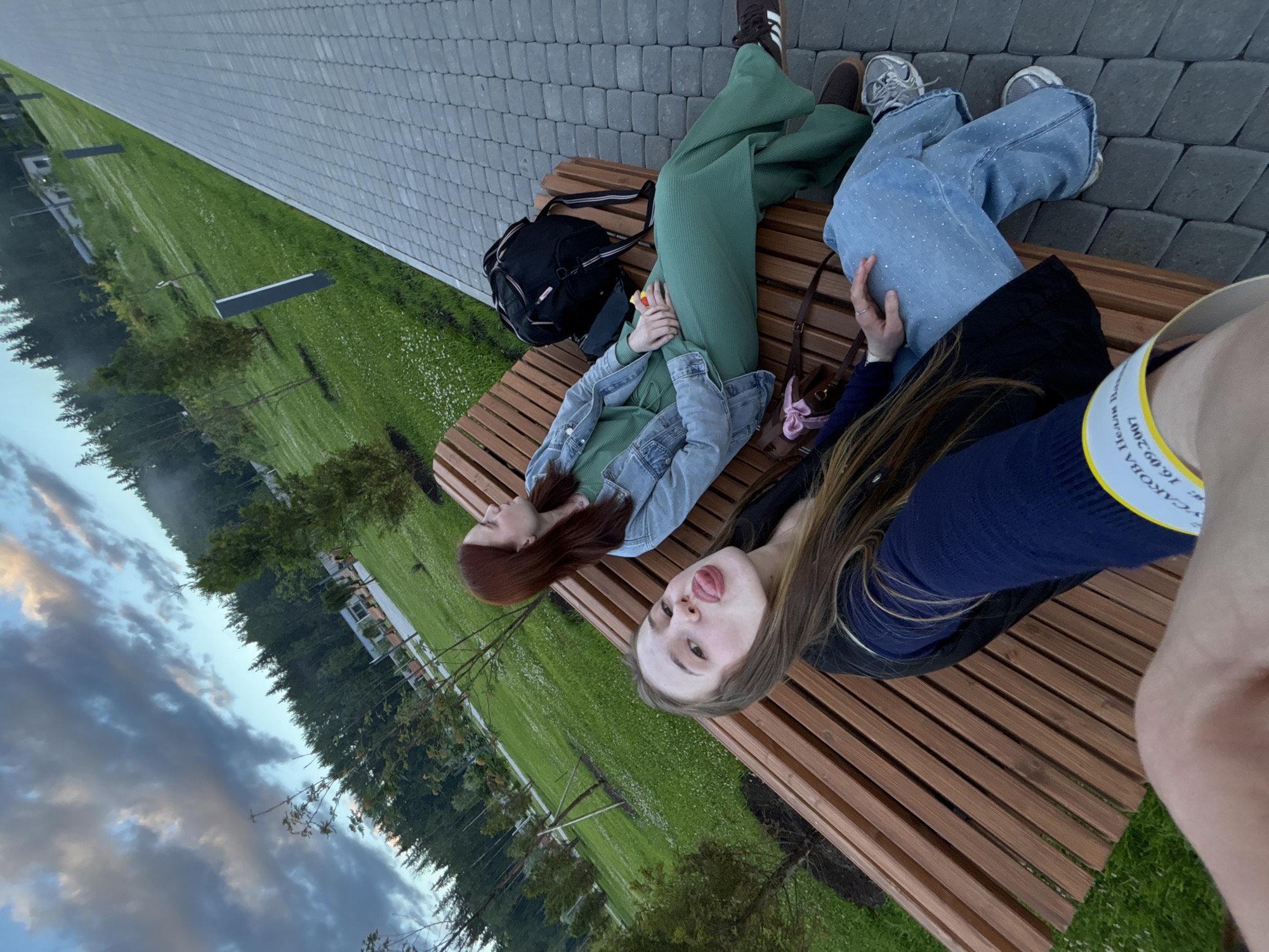 Two young women are seated on a wooden bench in a park. The woman in the foreground is facing the camera, sticking out her tongue. She is wearing a dark blue sweater and light blue jeans. The woman behind her is facing to the right, wearing a green jumpsuit and a denim jacket. A black backpack is on the bench behind her. The park features green grass, trees, and a paved walkway. The sky is cloudy with hints of sunset colors. The location is identified as Solnechnoye, Russia.