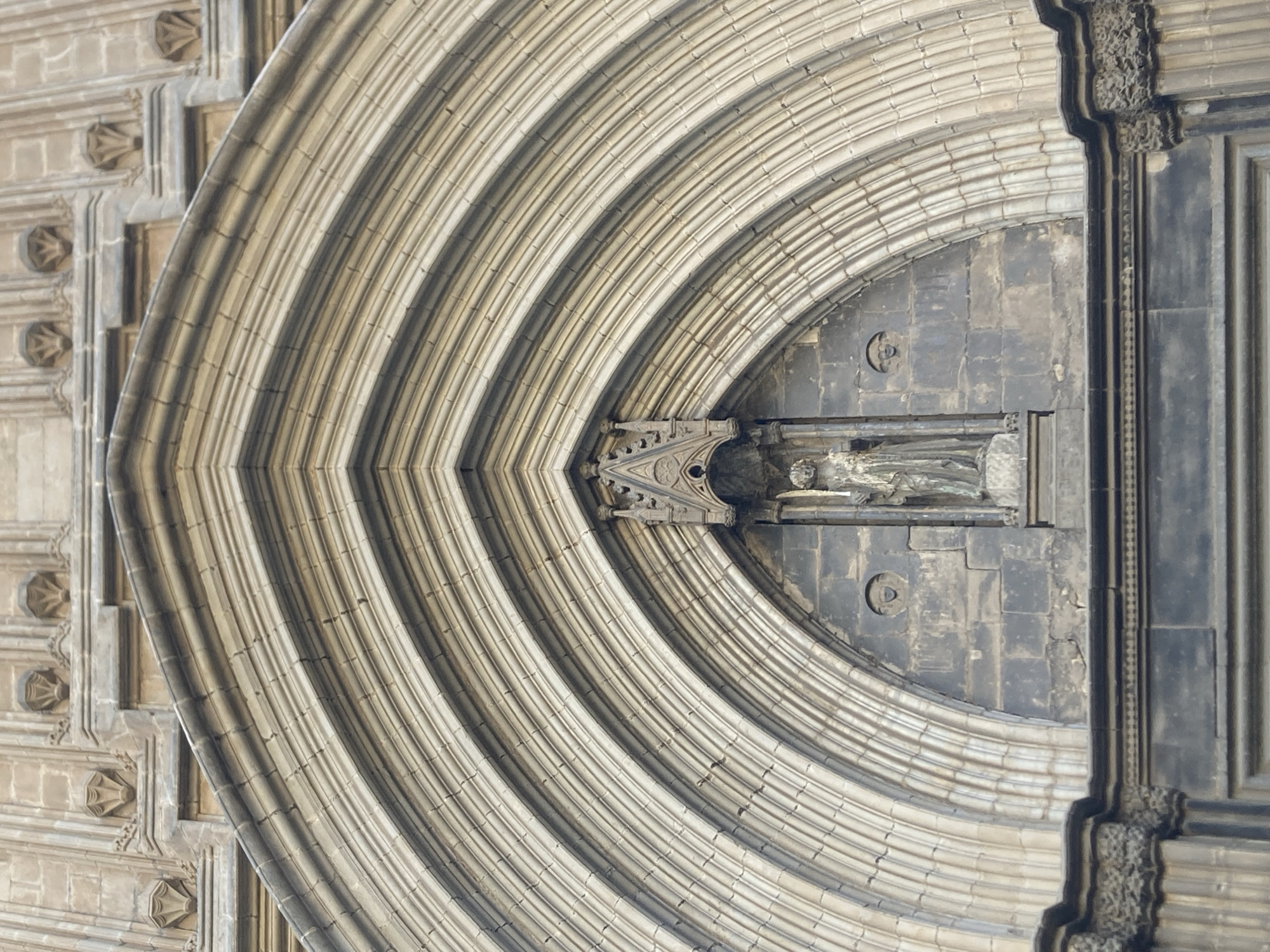 This media file shows a close-up, symmetrical view of a Gothic architectural detail, likely an archway or a facade element from a cathedral in Barcelona, Spain. The main subject is the intricate stonework, characterized by repeating, curved ribs that form a series of concentric arches. These ribs are textured and appear to be made of a light-colored stone, possibly sandstone or limestone, with darker shadowed lines between them creating a sense of depth and dimension.
Within the central part of the arched structure, there is a niche containing a sculpture, partially obscured. The sculpture appears to be of a figure, possibly religious in nature, carved from a darker material. The background consists of dark, roughly hewn stone blocks, providing a stark contrast to the lighter, more refined stonework of the arches.
The image is symmetrical along a vertical axis, suggesting it might be a reflection or a digitally manipulated composite of two halves. The lighting is soft and diffused, indicating it could be a cloudy day or the image was taken during a time when the sun was not directly hitting the facade, such as early morning or late afternoon. There are no people or active events depicted. The focus is solely on the architectural ornamentation, highlighting the craftsmanship and historical style. The overall impression is one of grandeur, antiquity, and detailed artistry. No visible text is present in the image.