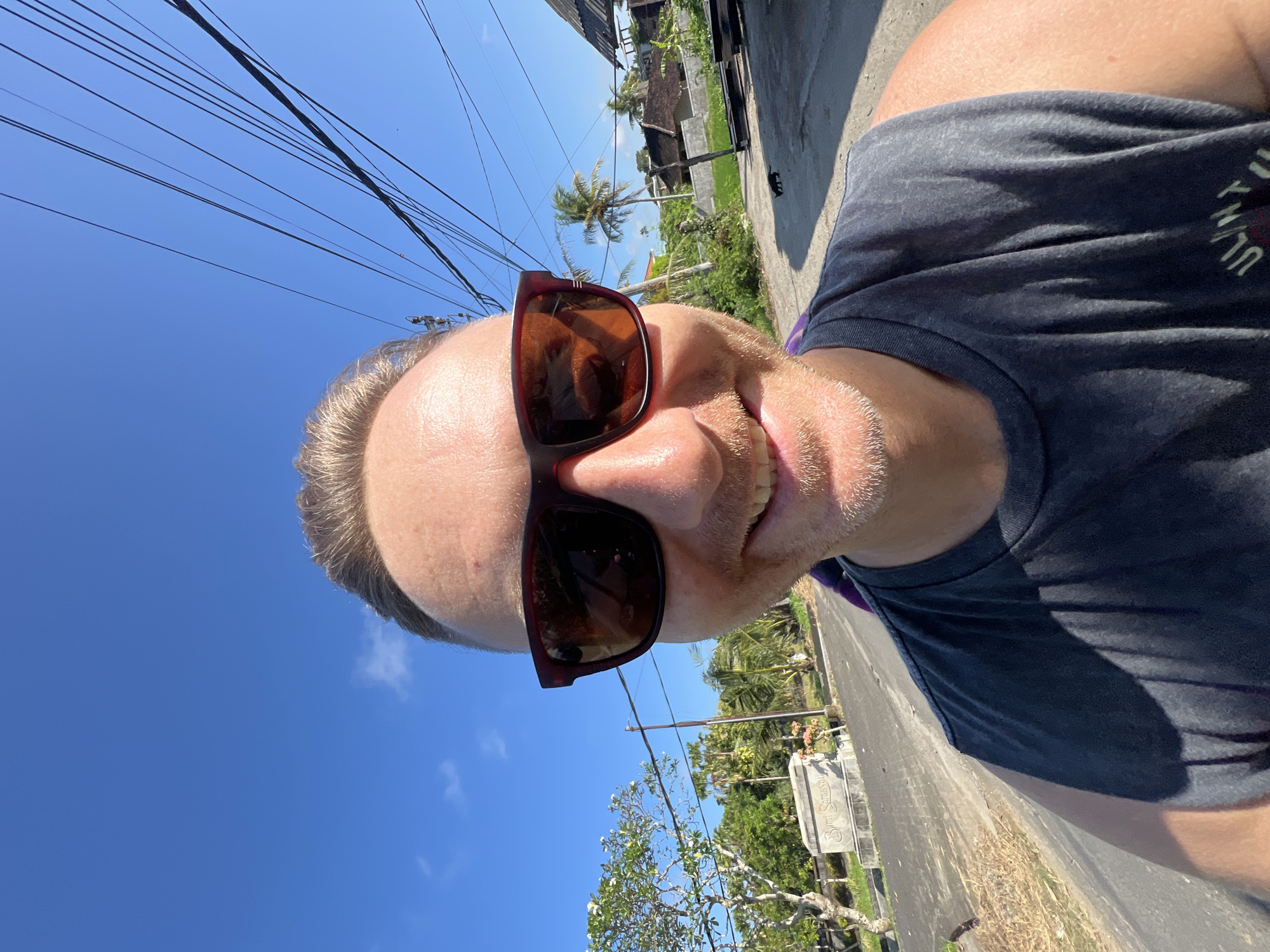 The image is a selfie of a light-skinned man wearing sunglasses and a dark gray t-shirt, taken outdoors in Sukawati, Indonesia. The man is positioned in the bottom right corner of the frame, with the camera pointed upwards at an angle. Overhead powerlines stretch diagonally across the top left of the image against a bright blue sky. In the background, a street with houses, greenery, and palm trees is visible. A white tombstone-like structure can be seen on the right. The sunlight is strong, casting shadows on the man's shirt and the road.