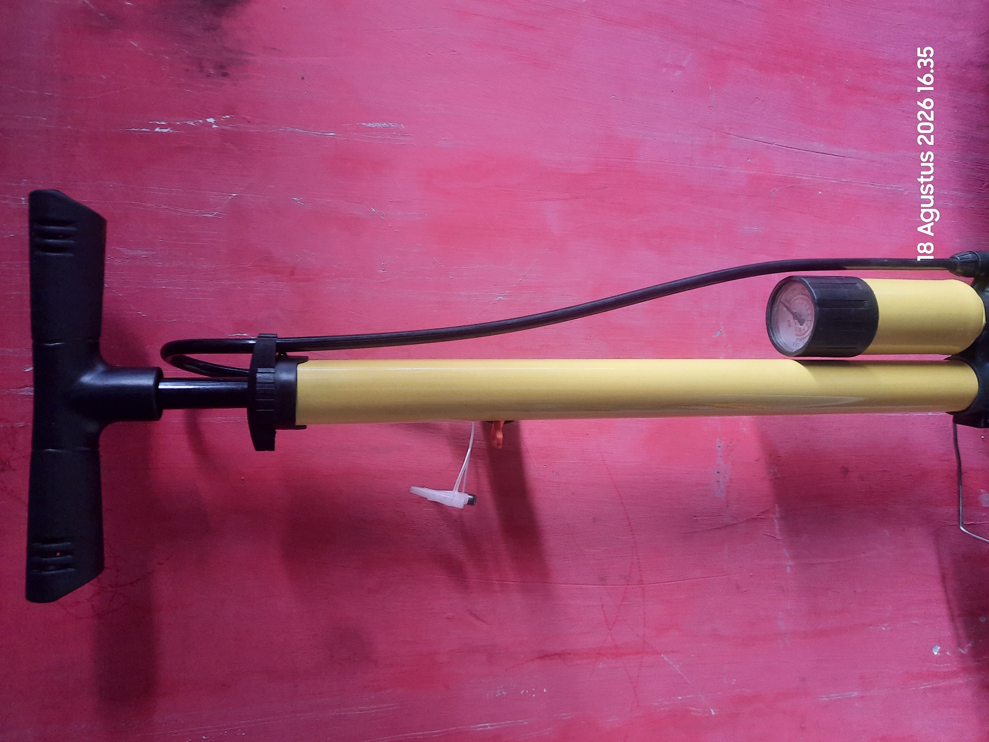 An upright, yellow bicycle pump with a black handle and a black hose is positioned against a textured, pink wall. The pump has a circular pressure gauge attached to its base, which is black and yellow. The handle of the pump is black and T-shaped, with horizontal indentations for grip. A white nozzle for inflating tires is attached to the pump by a black hose. The wall behind the pump shows signs of wear and tear, with visible scratches and imperfections. In the lower right corner of the image, text overlays the wall, reading "18 Agustus 2026 16.35". This text indicates a date and time, and implicitly suggests the location as Sukokerto, Indonesia. There are no people or animals visible in the image, and no actions or interactions are depicted. The lighting suggests an indoor or shaded outdoor environment.