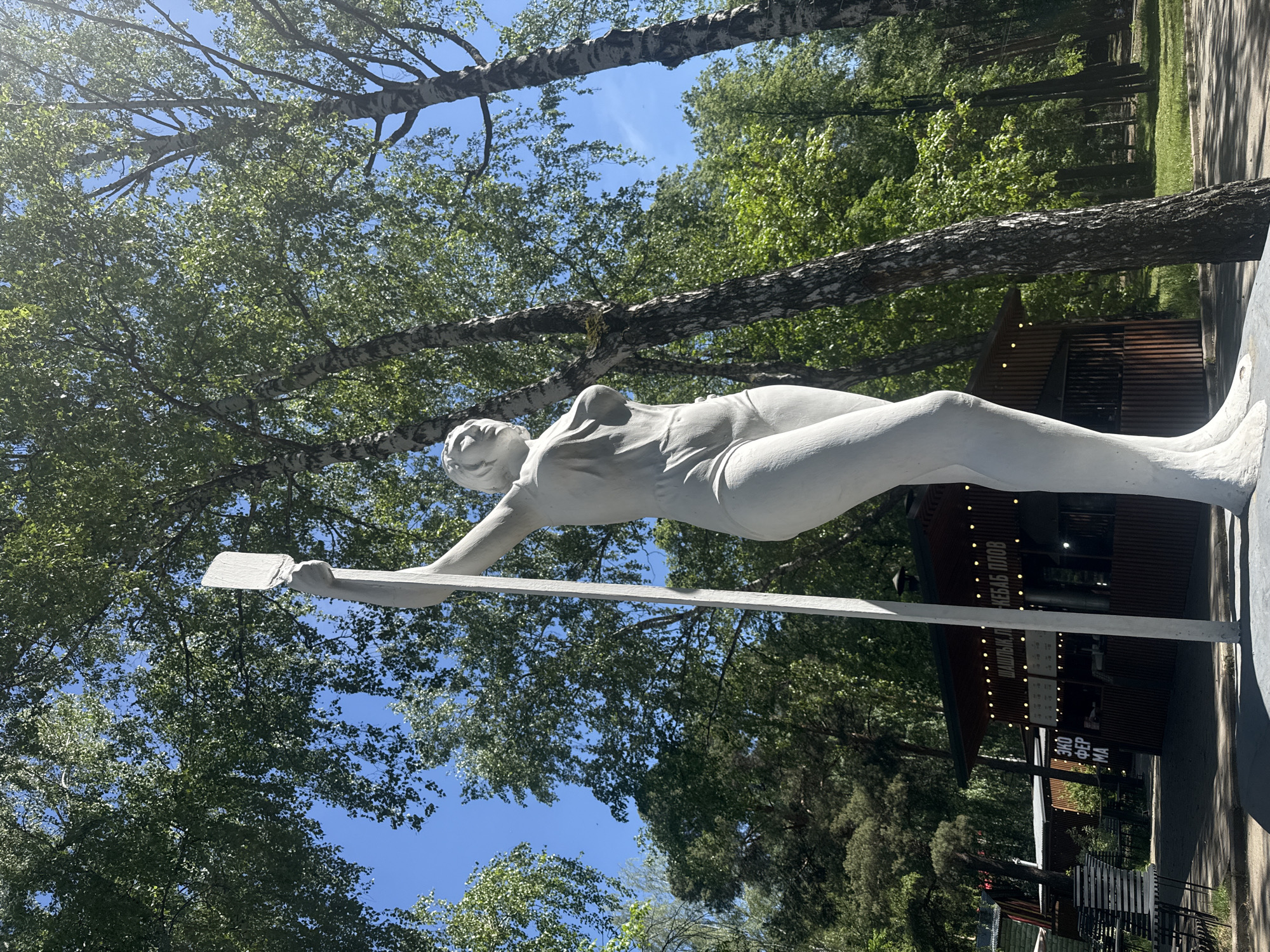 The image shows a white statue of a rower in Yekaterinburg, Russia, lying horizontally with an oar in her hands. The statue is positioned against a backdrop of green trees and a blue sky. A small building with lights and text in Cyrillic is visible behind the statue.