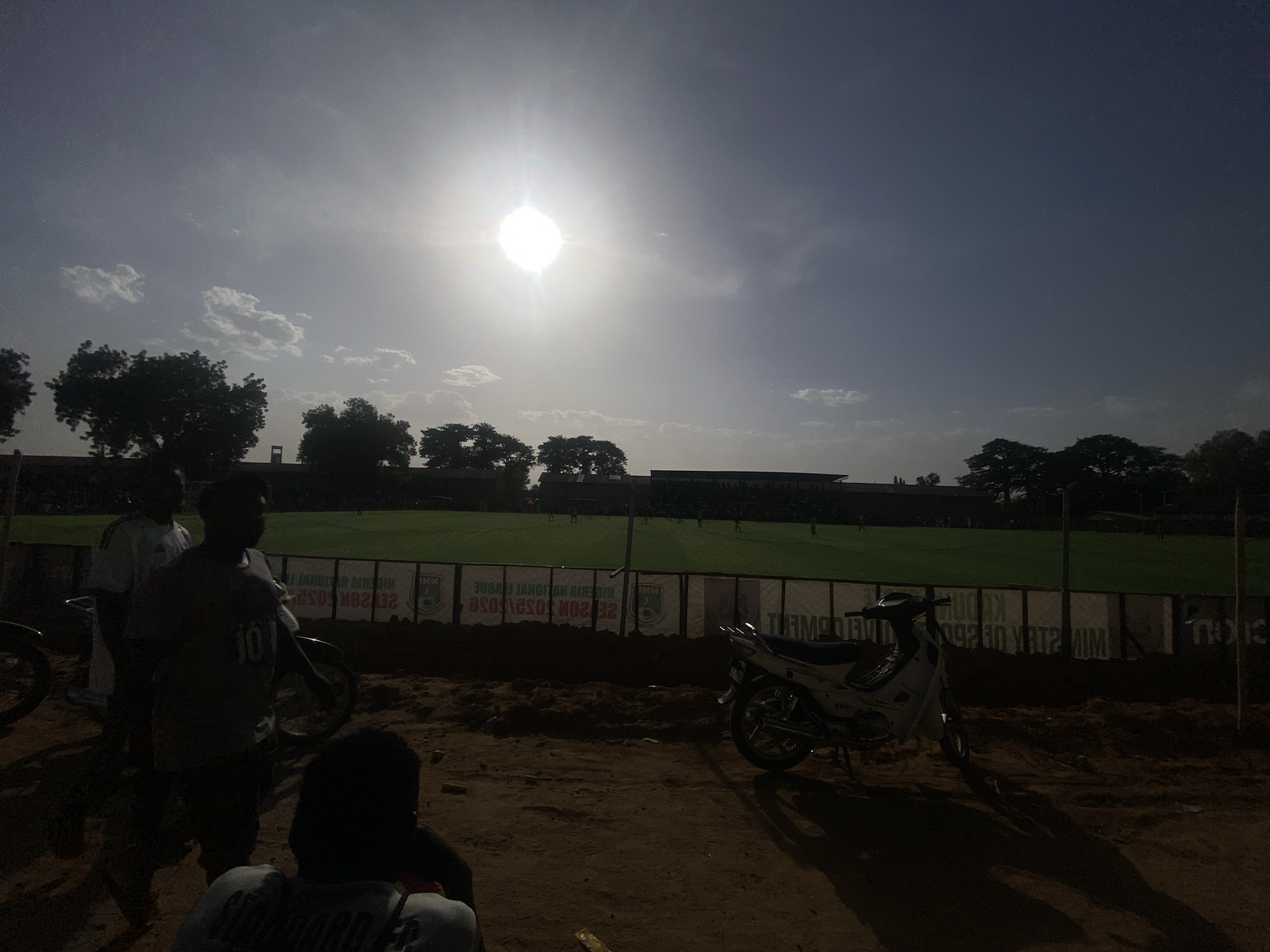 The image shows a daytime scene in Tudun Wada, Nigeria. A football field is visible in the background, surrounded by a fence and trees. Several people are standing and sitting in the foreground, along with two motorcycles parked nearby. The sun is high in the sky, causing a bright glare. The ground appears to be dirt or sand.
