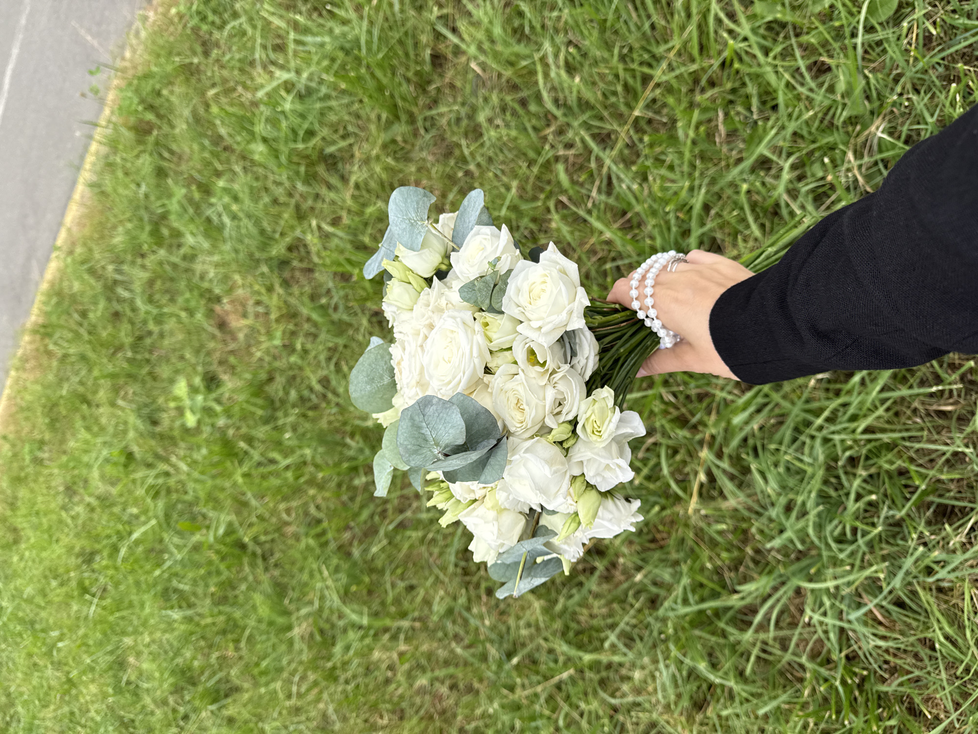 A hand wearing a black long-sleeved shirt and two silver rings, adorned with a pearl bracelet, holds a bouquet of white roses and eucalyptus branches. The bouquet is presented against a backdrop of lush green grass, bordering a paved surface, suggesting an outdoor urban setting, possibly Votkinsk, Russia.
