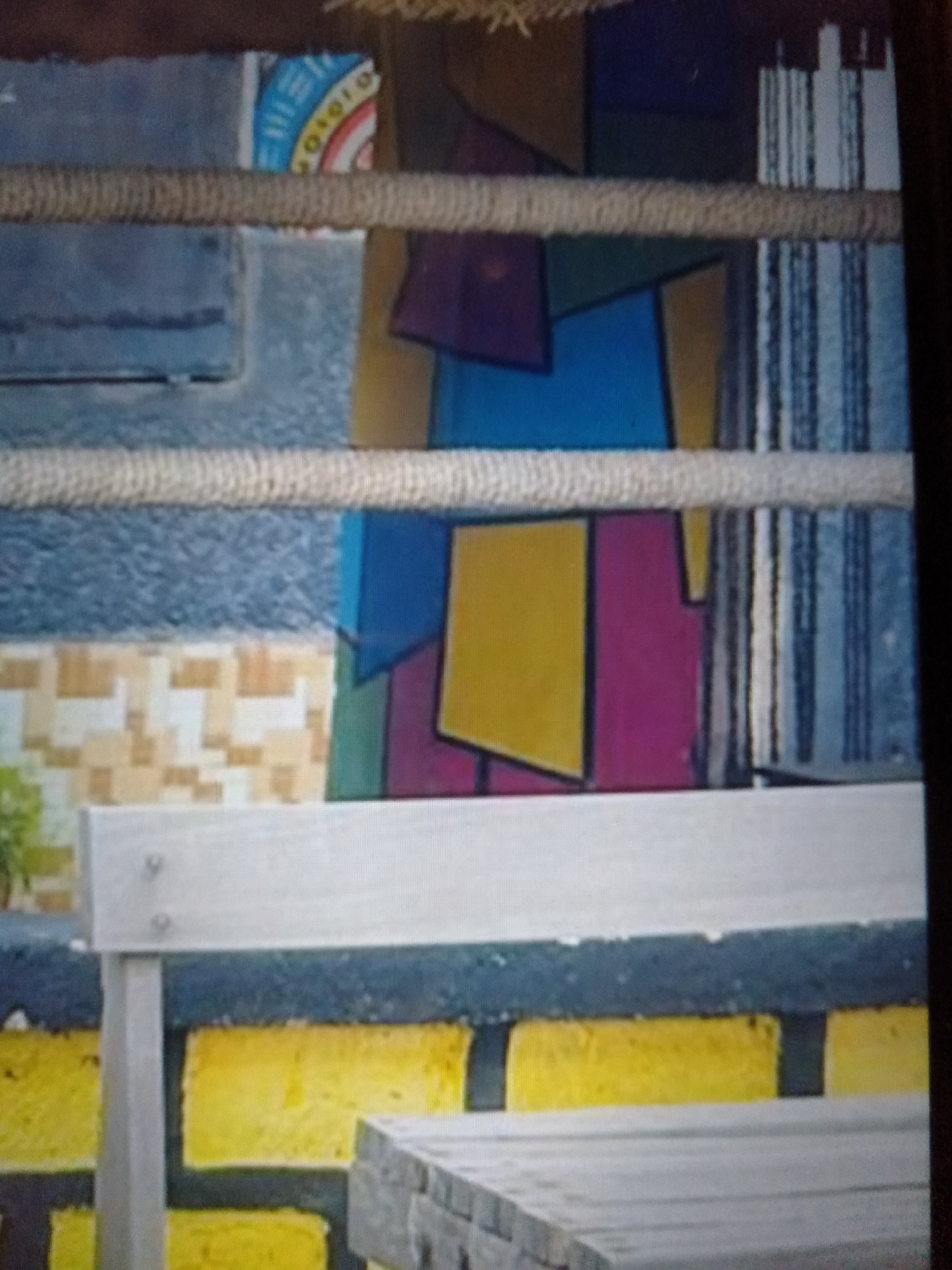 The image shows an outdoor setting, possibly a seating area or a decorative wall, with a strong emphasis on geometric patterns and vibrant colors. In the foreground, a light-colored bench with metal supports is visible, placed in front of a wall constructed of yellow and dark gray rectangular blocks. To the right of the bench, a portion of a mosaic-tiled tabletop or surface is also present.
The background is more abstract and colorful. It features a series of irregularly shaped blocks of color, including shades of blue, yellow, pink, and green, arranged in a stained-glass or mosaic-like fashion. Above these colorful blocks, there are striped patterns in blues and grays, and to the left, a textured gray surface with a section of a circular design containing a rainbow and some blue and white text. A thick, rope-like material stretches horizontally across the middle of the image, obscuring parts of the background. The lighting suggests it is daytime. While Jalingo, Nigeria is provided as a location context, there are no explicit visual cues within the image to confirm this specific location. No discernible emotion is conveyed.