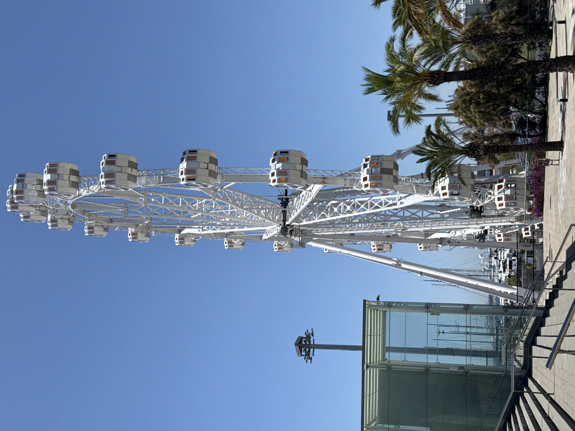 The image shows a tall, white Ferris wheel situated in a clear blue sky. The Ferris wheel has multiple enclosed cabins with orange-tinted windows. In the foreground to the left, there is a modern glass building with stairs leading up to it. A street lamp with multiple lights is visible behind the glass building. Palm trees are scattered throughout the scene, some in the foreground and others in the mid-ground. In the background, a body of water with numerous sailboats and yachts can be seen, suggesting a marina setting. This Ferris wheel is likely the "Rueda de Barcelona" or "Port Olímpic Ferris Wheel," located in the Port Olímpic area of Barcelona, Spain. No people are visible in the image.