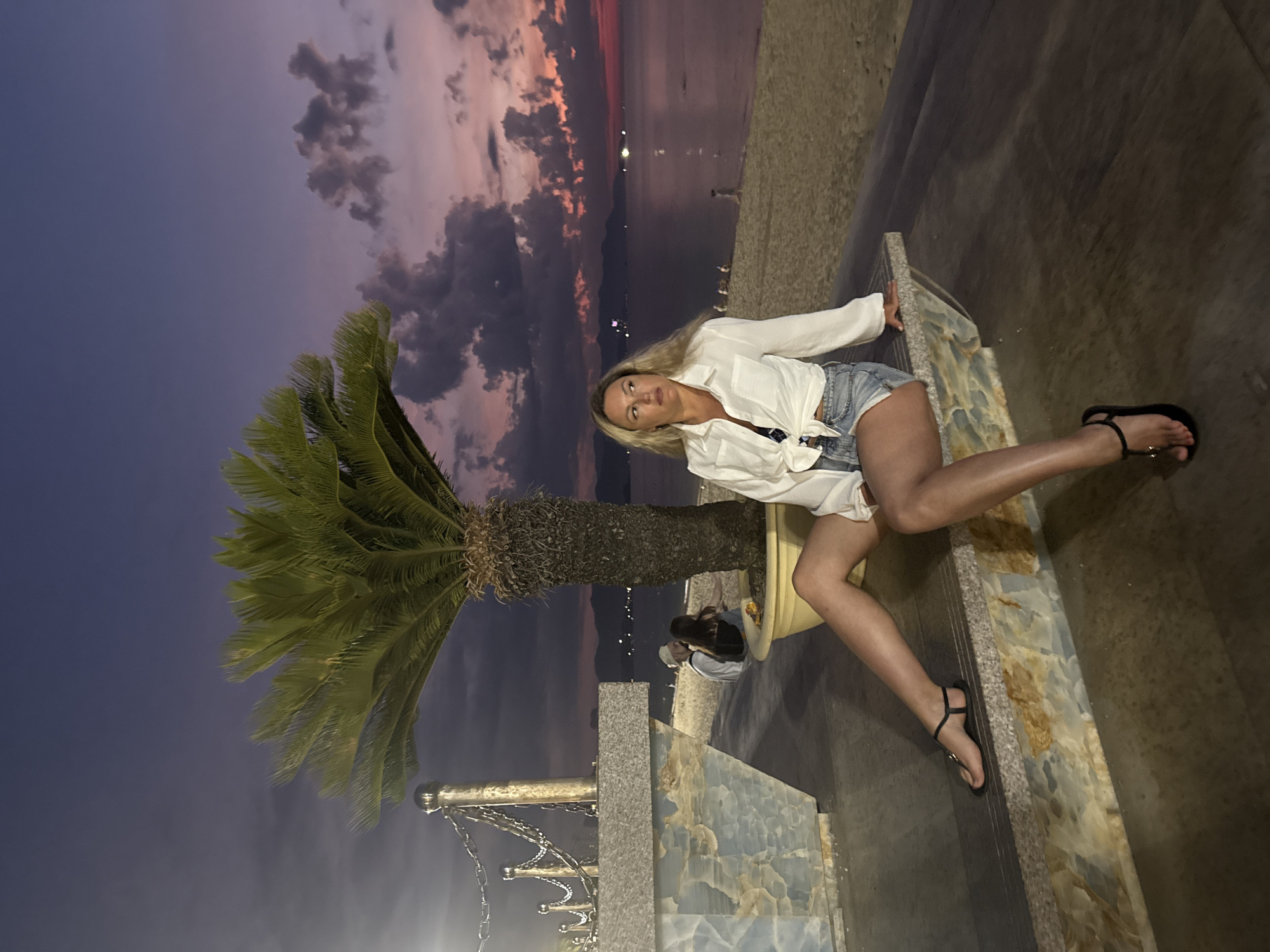 A woman with blonde hair is posing on a concrete ledge near a palm tree, with the sky and the sea visible in the background, likely in Nha Trang Ward, Vietnam. She is wearing a white shirt and denim shorts and black sandals. The sky displays colors from dusk and dawn. A couple is visible in the background by the tree. There is ornamental railing in the lower left of the frame. The image is rotated counter-clockwise.