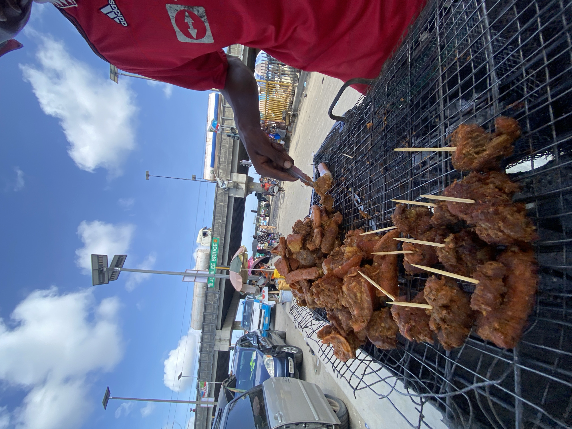 A street vendor grills skewered meat, potentially suya, at a roadside grill. The location is identified as Lekki, Nigeria, as indicated by the "Jubilee Bridge" sign visible in the background. The vendor, wearing a red shirt with Adidas and what appears to be an insurance company logo, uses tongs to manipulate the meat on the grill. Cars are parked nearby, with a blue sky and scattered clouds in the background.