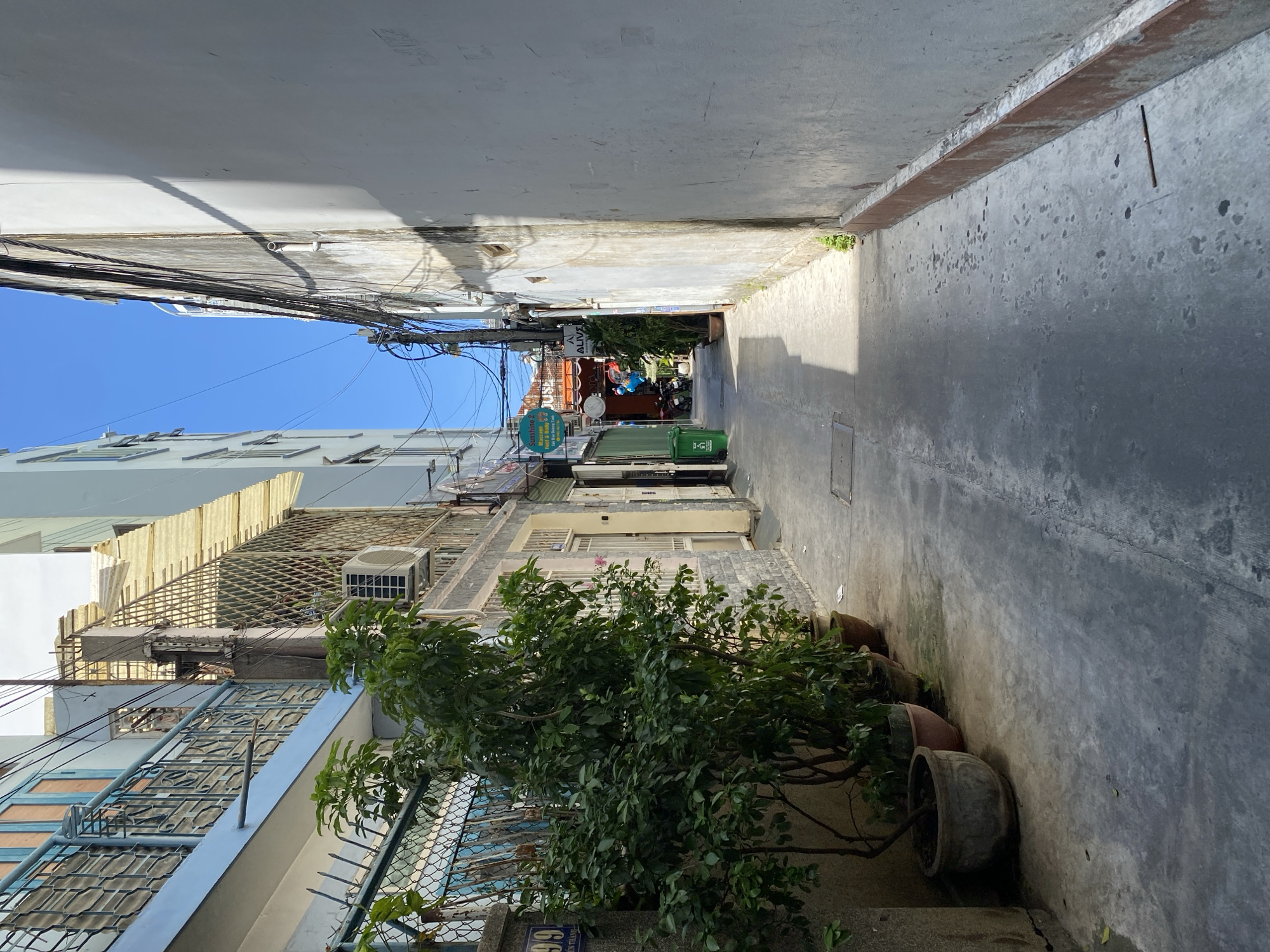 The image depicts a narrow alleyway in Nha Trang Ward, Vietnam, flanked by buildings on either side. The alley's ground is paved with concrete, showing signs of wear and scattered water stains. On the left, a building with a light blue facade features balconies with ornate metal railings and wooden-framed windows. Several potted plants, including a large leafy green one, are placed along the base of this building. Further down the alley on the left, a green trash bin is visible. The right side of the alley is dominated by a tall, weathered white wall with a reddish-brown trim at its base. The wall casts a long shadow from an unseen object. Overhead, a tangle of electrical wires crisscrosses the sky, which is a clear blue. In the distance, signs for businesses like "Sunshine Massage" and "HOUSE" are visible, and a few motorbikes are parked. The overall impression is that of a bustling, lived-in urban environment.