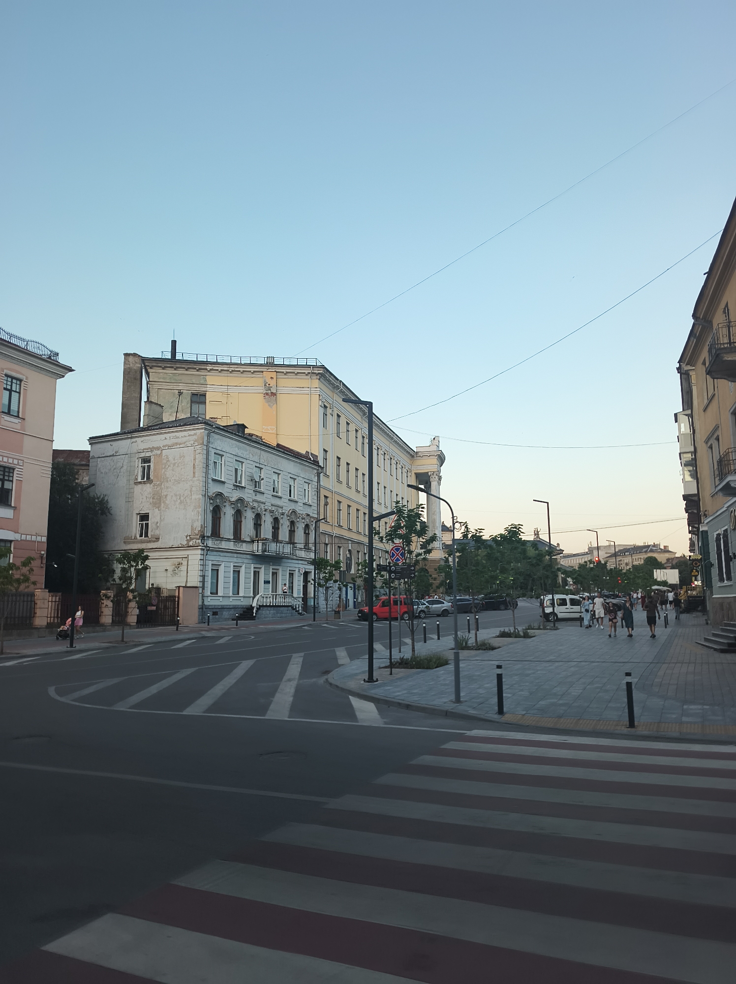 The image depicts a street scene in Ternopil, Ukraine, with historical buildings lining the right side of the thoroughfare. The buildings feature architectural details typical of earlier centuries, including ornate windows and balconies. On the left, a lighter-colored building is visible, also with multiple stories and windows. The street itself is paved and has clear pedestrian crosswalks marked with white and red stripes.
A sidewalk runs along the right side of the street, populated by several individuals walking. The people are dressed in casual summer clothing. Some are walking in groups, while others appear to be alone. A few trees are planted along the sidewalk, providing some greenery.
Vehicles are present on the street, including cars and a white van. Traffic appears to be moving, though not densely. Overhead, power lines are strung across the sky, a common sight in urban environments. The sky is clear and a pale blue, indicating daylight, likely late afternoon or early evening given the soft lighting. The overall atmosphere is calm and ordinary, capturing a typical urban street in Ukraine.