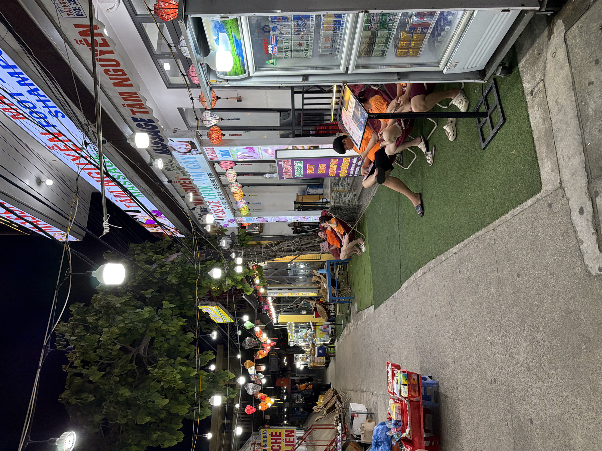 The image depicts a nighttime scene on a street in Da Nang, Vietnam, likely a commercial area. The foreground is dominated by a concrete walkway with several red plastic baskets filled with assorted goods, possibly snacks or small retail items, placed on a red cart. To the right, a section of artificial turf lawn is visible, where several individuals are seated on lounge chairs.
A white commercial refrigerator, stocked with canned beverages, stands prominently on the right side of the frame. Above the lawn area, a building facade displays multiple signs in Vietnamese, advertising "MASSAGE TAM NGUYEN," "MASSAGE FOOT," and "SHAMPOO." Red and white lanterns are strung across the street, illuminating the area, along with several white globe-shaped lights. Trees with dense green foliage hang over the scene from the left, with their branches intertwined with the light strings.
In the middle ground, a man and a woman, both wearing orange shirts, are seated on lounge chairs facing a small table with what appears to be reading material. Another person, also in an orange shirt, is seated in a lounge chair further back on the right. The background shows more of the street with illuminated signs, food stalls, and other commercial establishments, suggesting a lively and bustling environment. The overall impression is that of a street market or a commercial strip offering services and goods, likely catering to both locals and tourists.